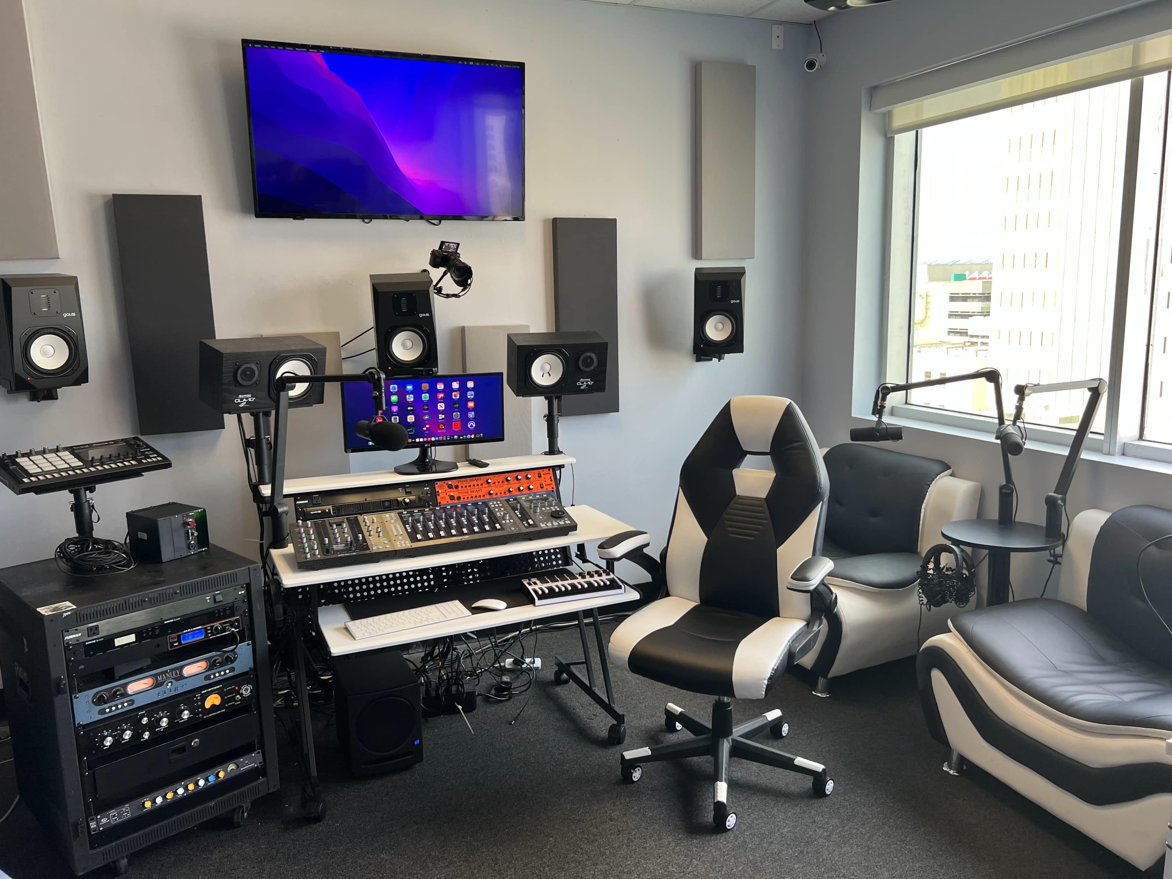 A music studio with audio equipment, including speakers, a mixing console, and a large computer monitor, complemented by ergonomic office chairs.