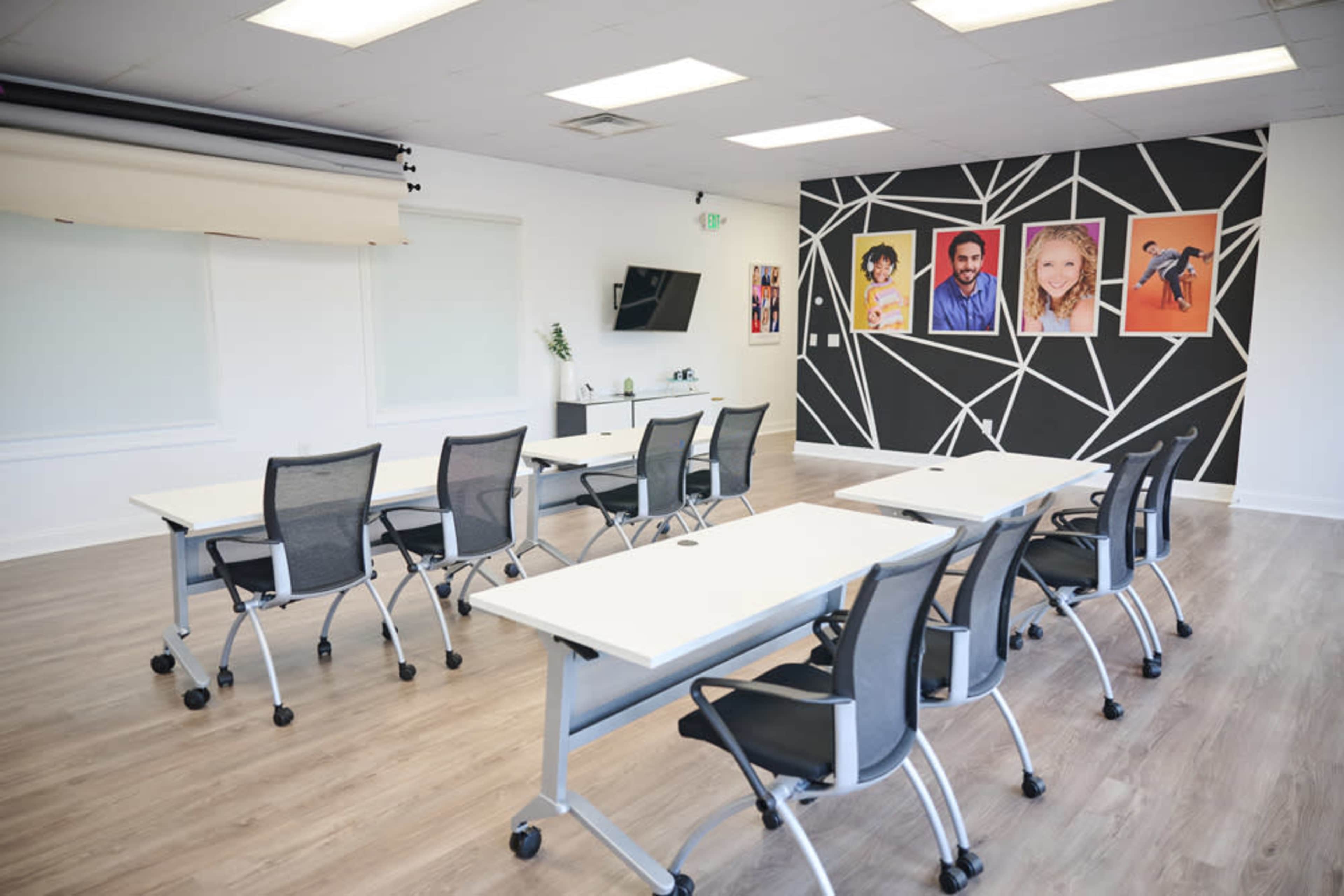 The image shows a modern classroom or meeting space featuring several tables and chairs arranged in a structured layout, with a black and white geometric mural and framed portraits on the wall.