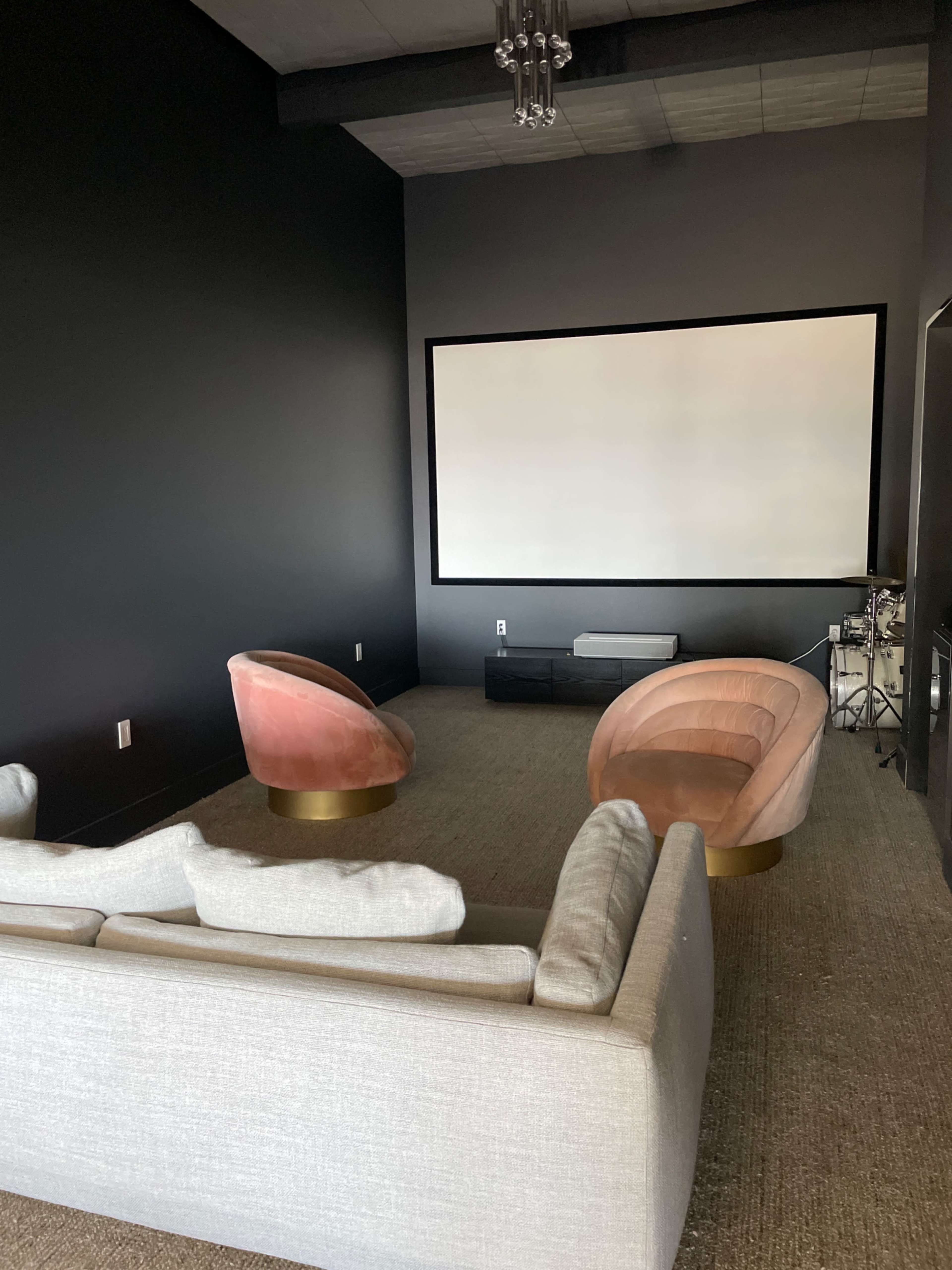 A modern lounge area features two accent chairs and a sofa, facing a large blank screen against a dark wall.