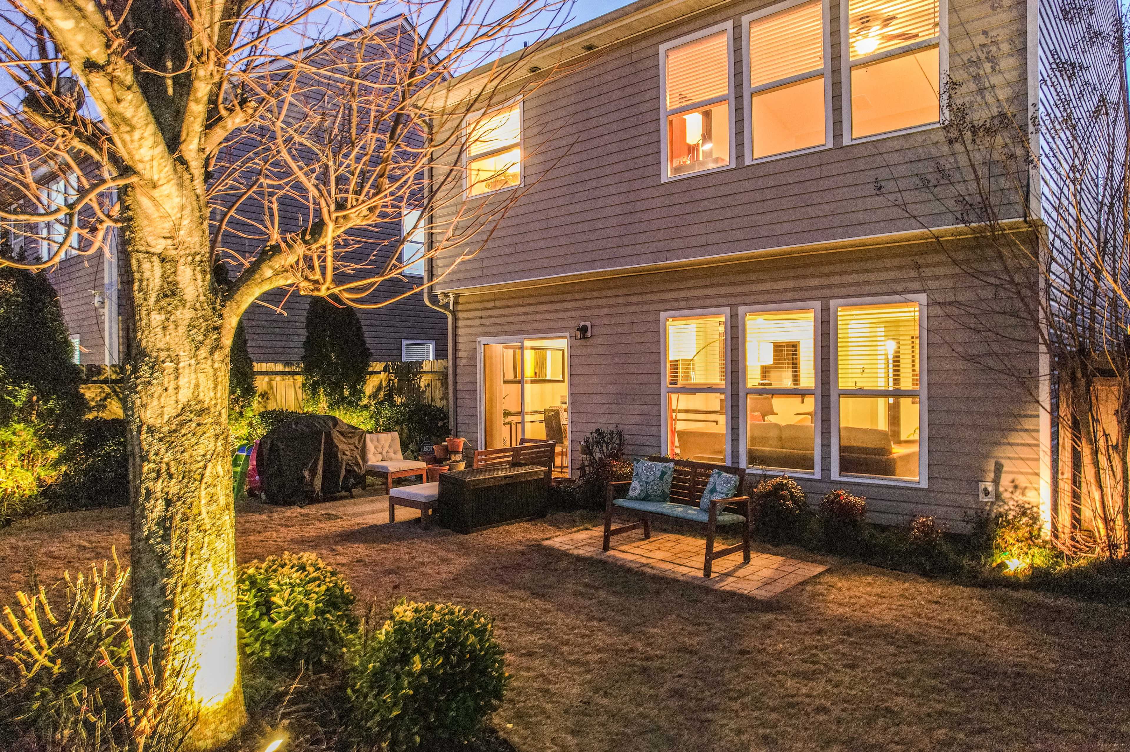 A two-story house with large windows is seen from the backyard at dusk, featuring a wooden patio, outdoor seating, and landscaped plants.