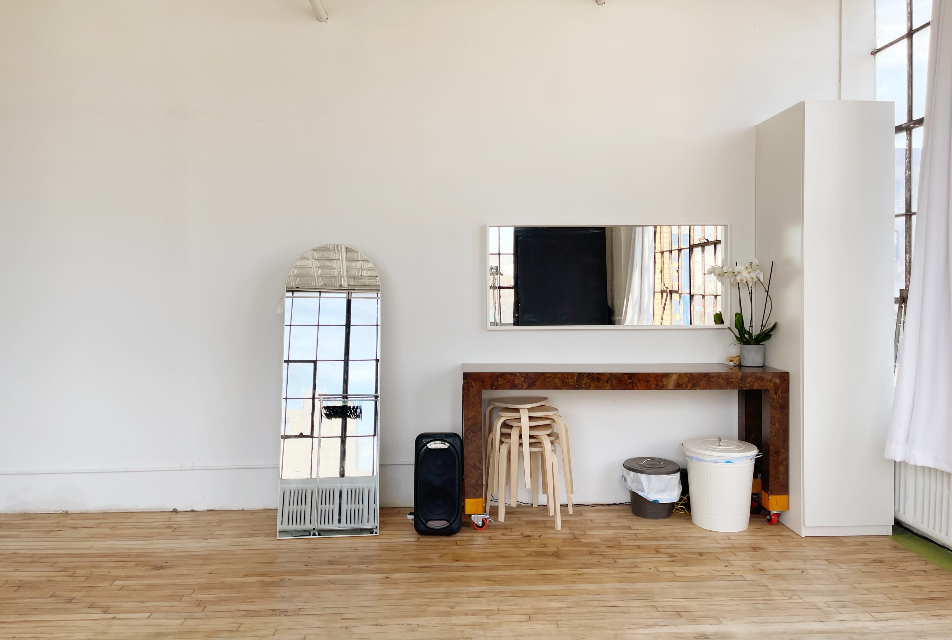 A minimalist interior features a wooden table with stools, a large mirror, a trash can, and a speaker against a white wall with windows.