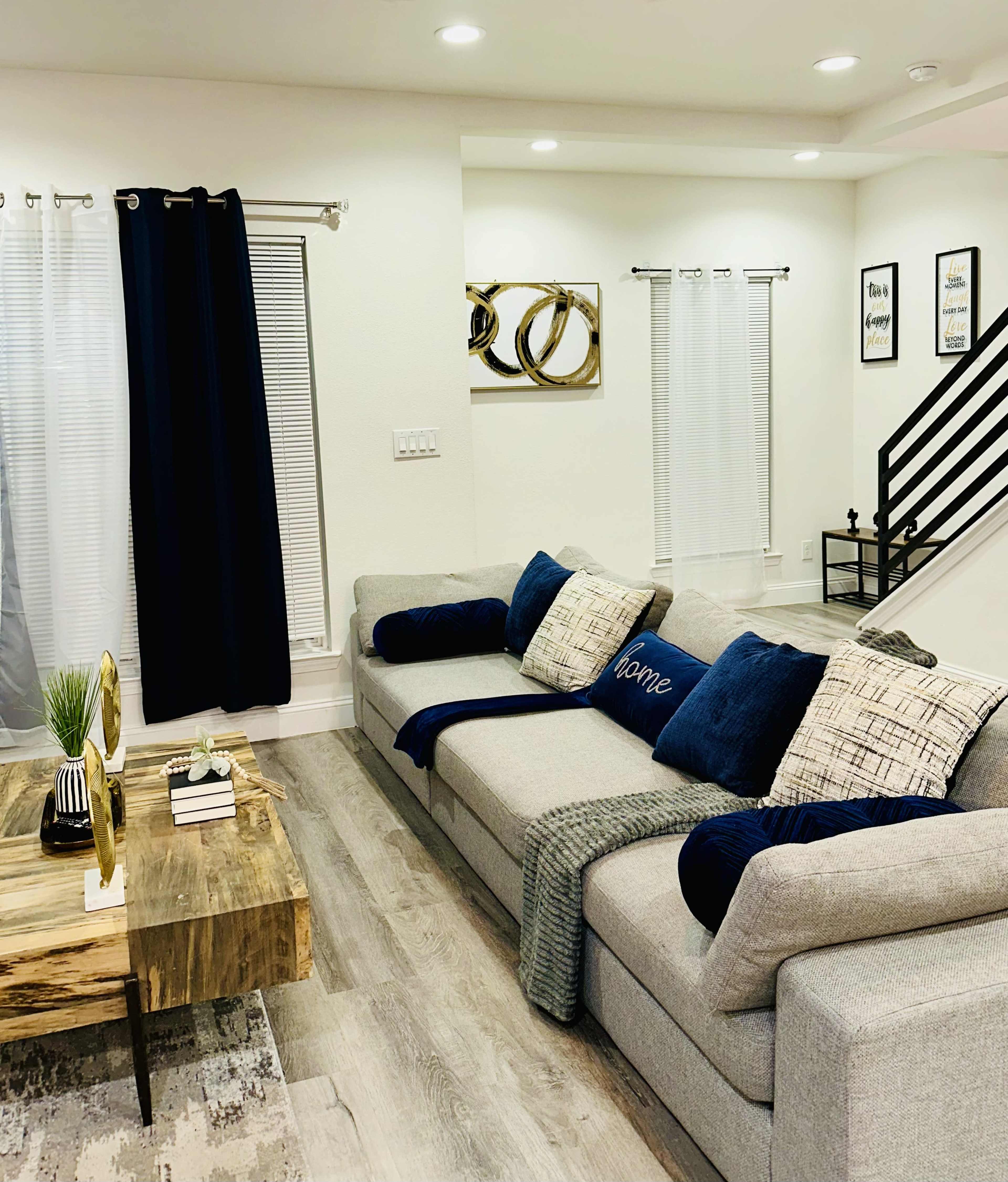 A modern living room featuring a gray sectional sofa with blue and white throw pillows, a wooden coffee table, and framed wall art, with light-colored walls and hardwood flooring.