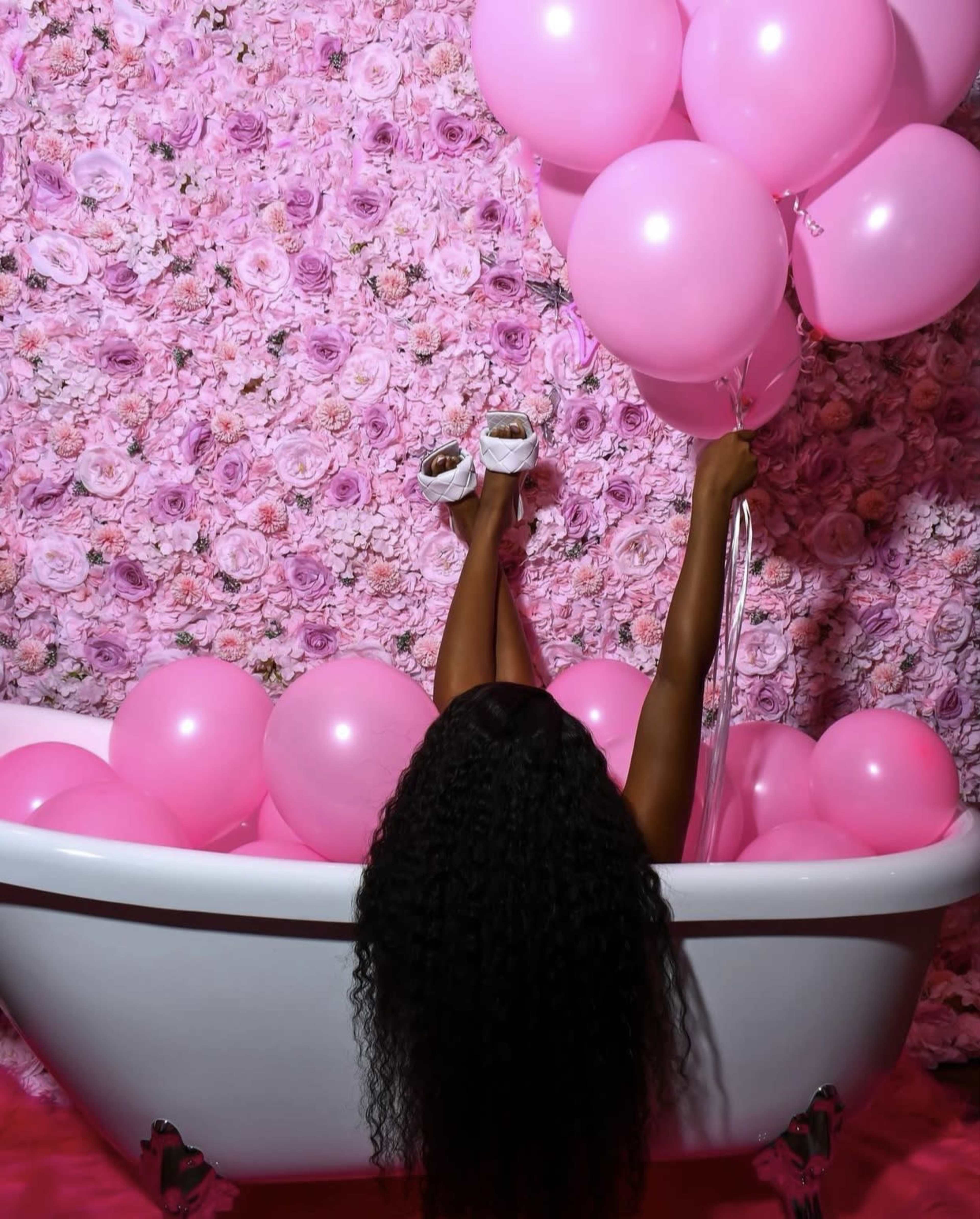 A person relaxes in a white bathtub filled with pink balloons against a backdrop of a floral pink wall.