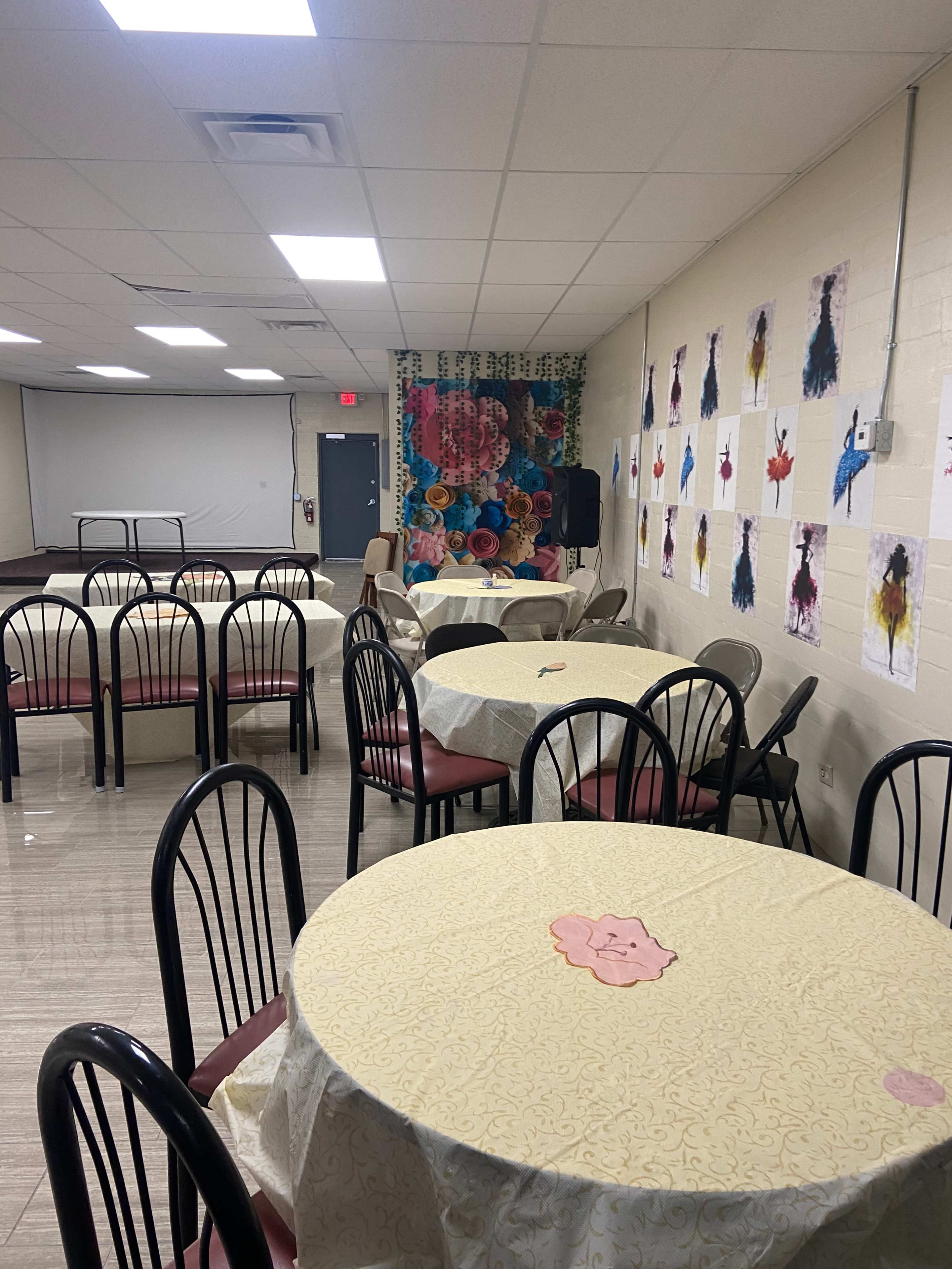 A spacious indoor room set up with round tables covered in light-colored tablecloths, accompanied by chairs and decorated walls featuring various artworks.