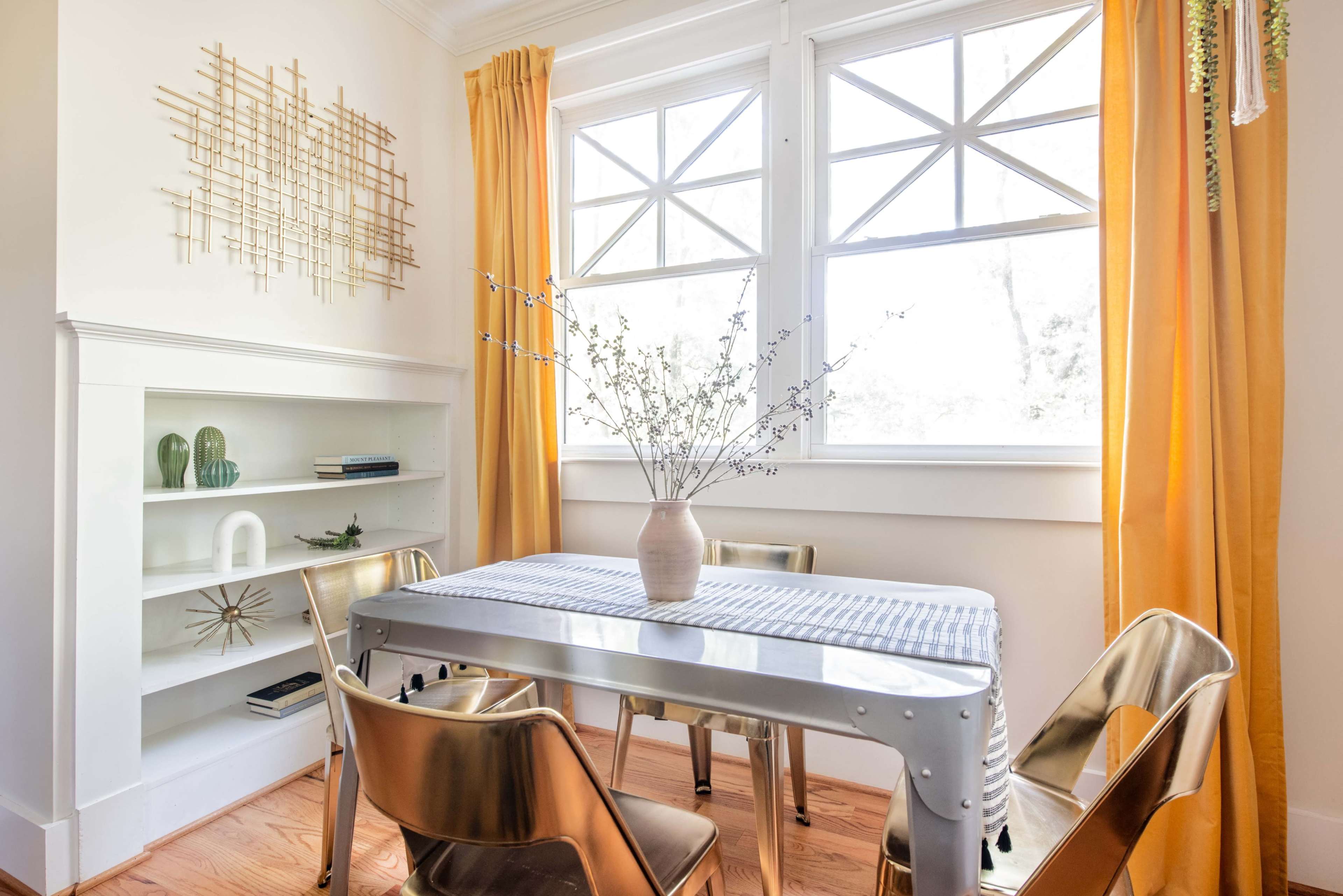 A dining area features a metal table surrounded by four chairs, with a vase of branches on the table and large windows draped with yellow curtains.