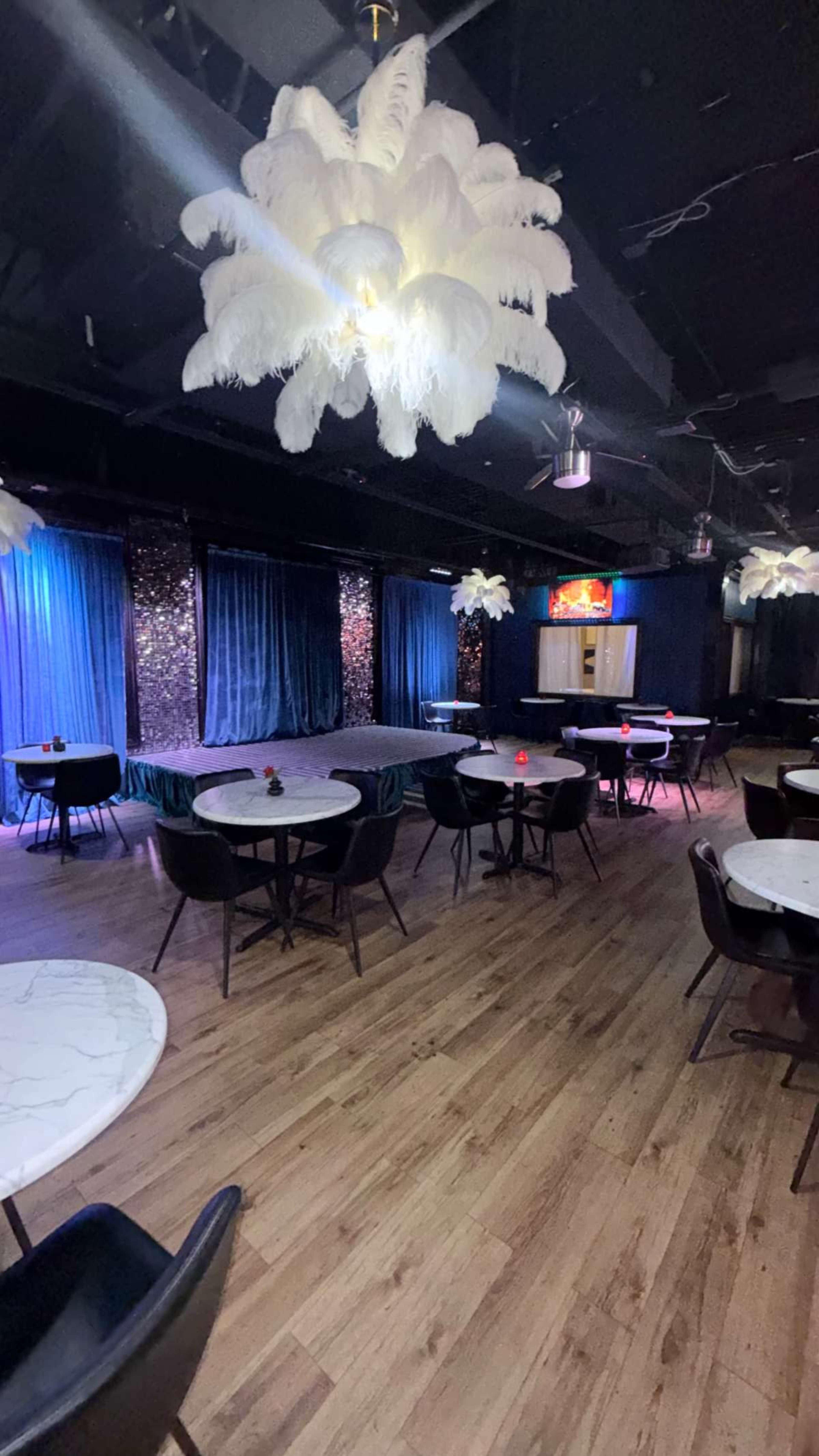The image shows an indoor venue with a stage, tables, and decorative lighting featuring large feather-like chandeliers.