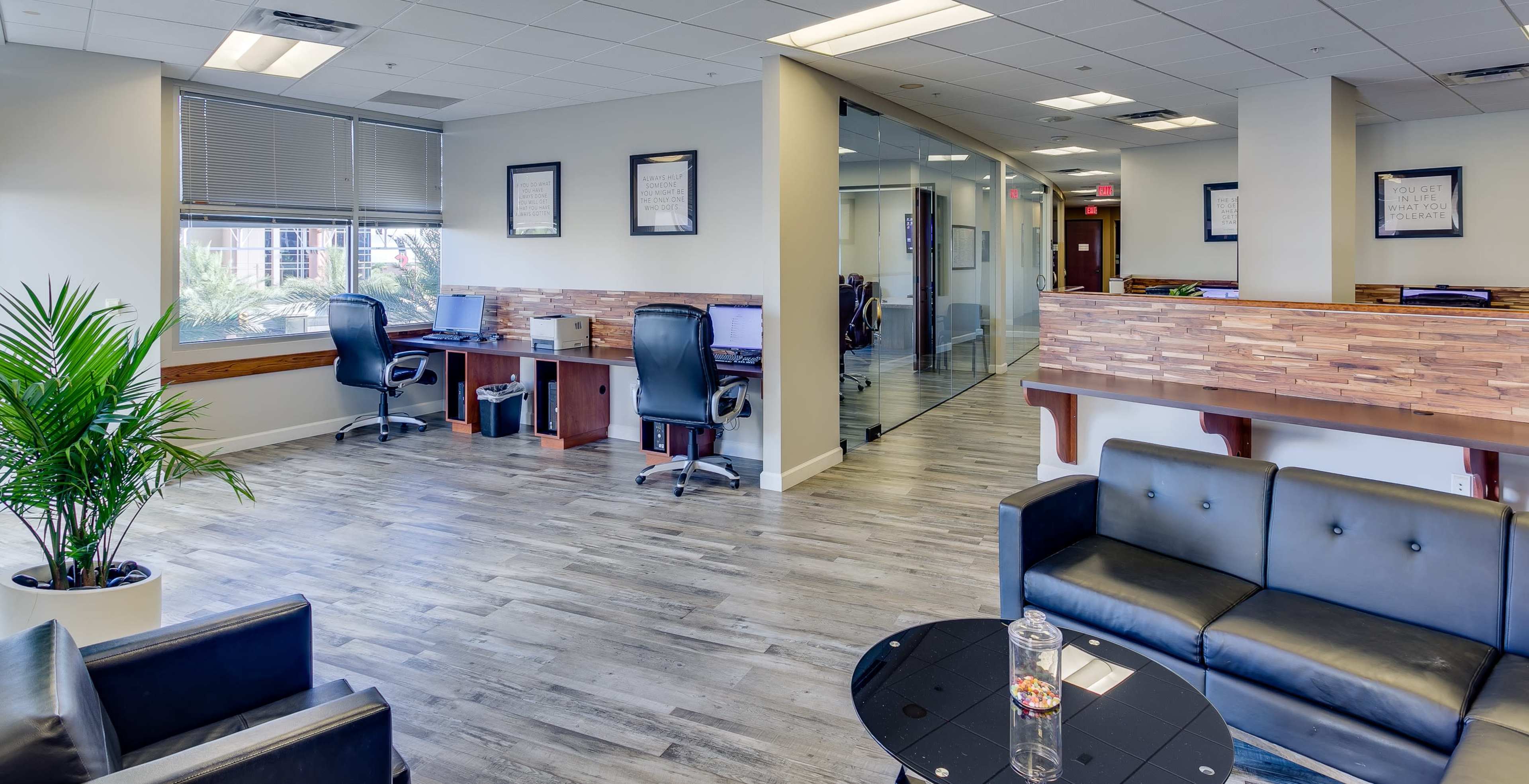 An office space featuring two desks with chairs, a seating area with a black couch and a glass table, and a corridor leading to additional rooms.