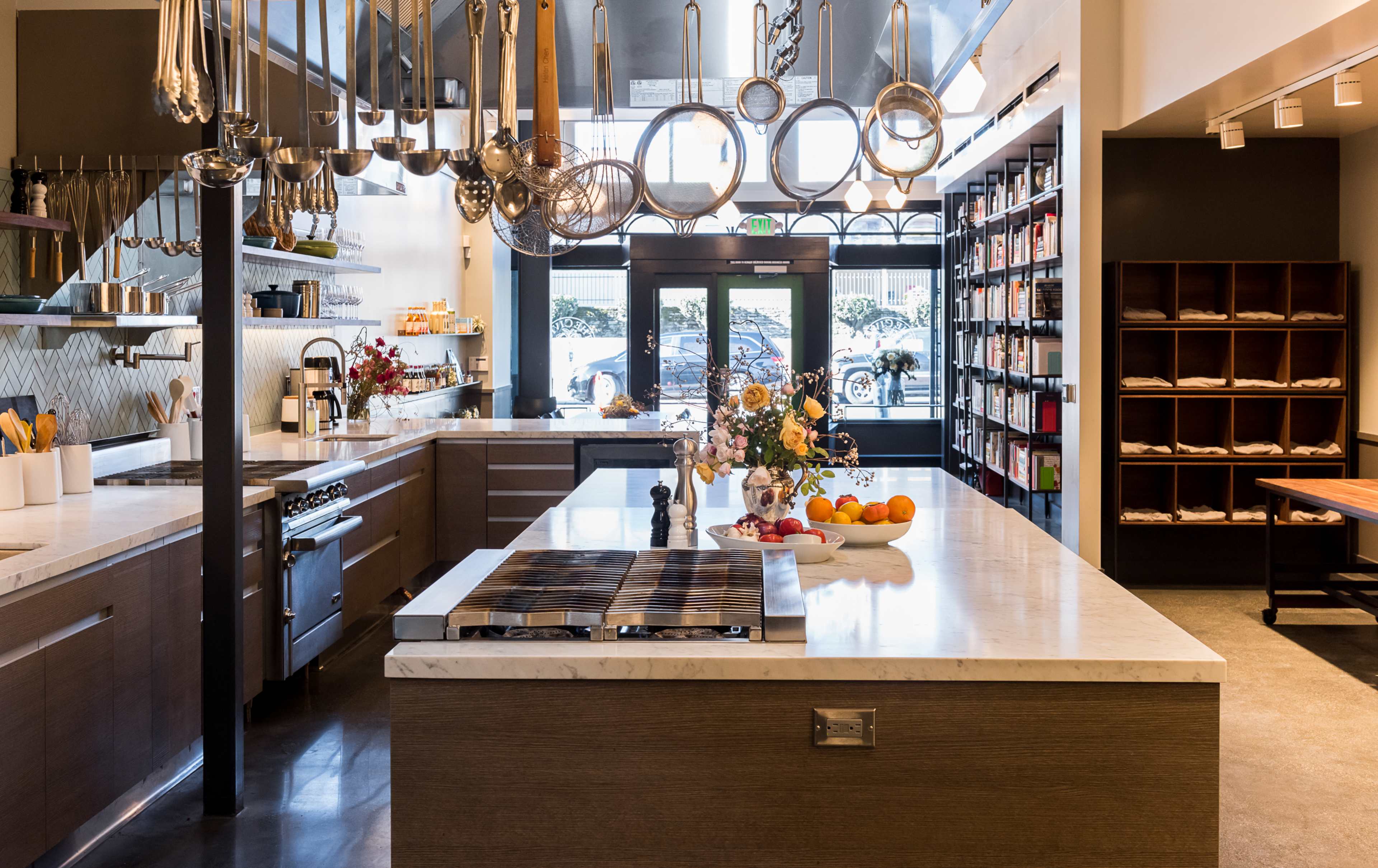 A modern kitchen features a spacious island with a stovetop, hanging pots and utensils, and shelves filled with various kitchen supplies.