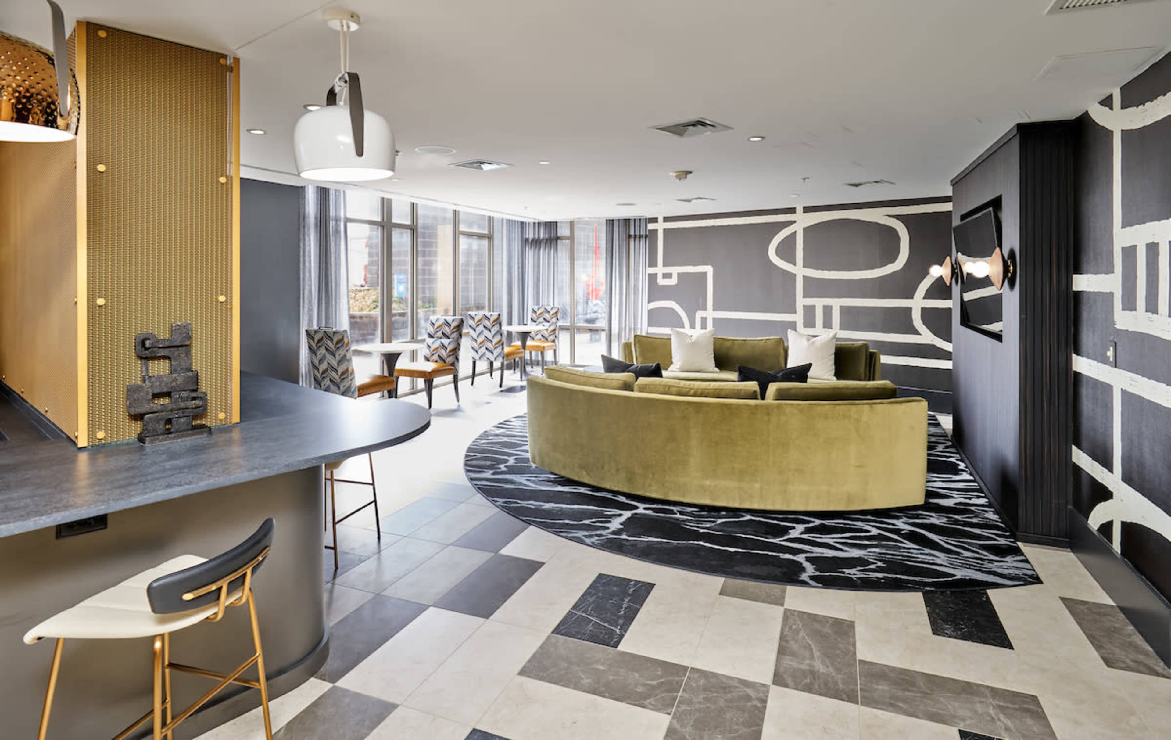 A modern lounge area featuring a curved green sofa, patterned chairs, and a geometric rug, with large windows providing natural light.