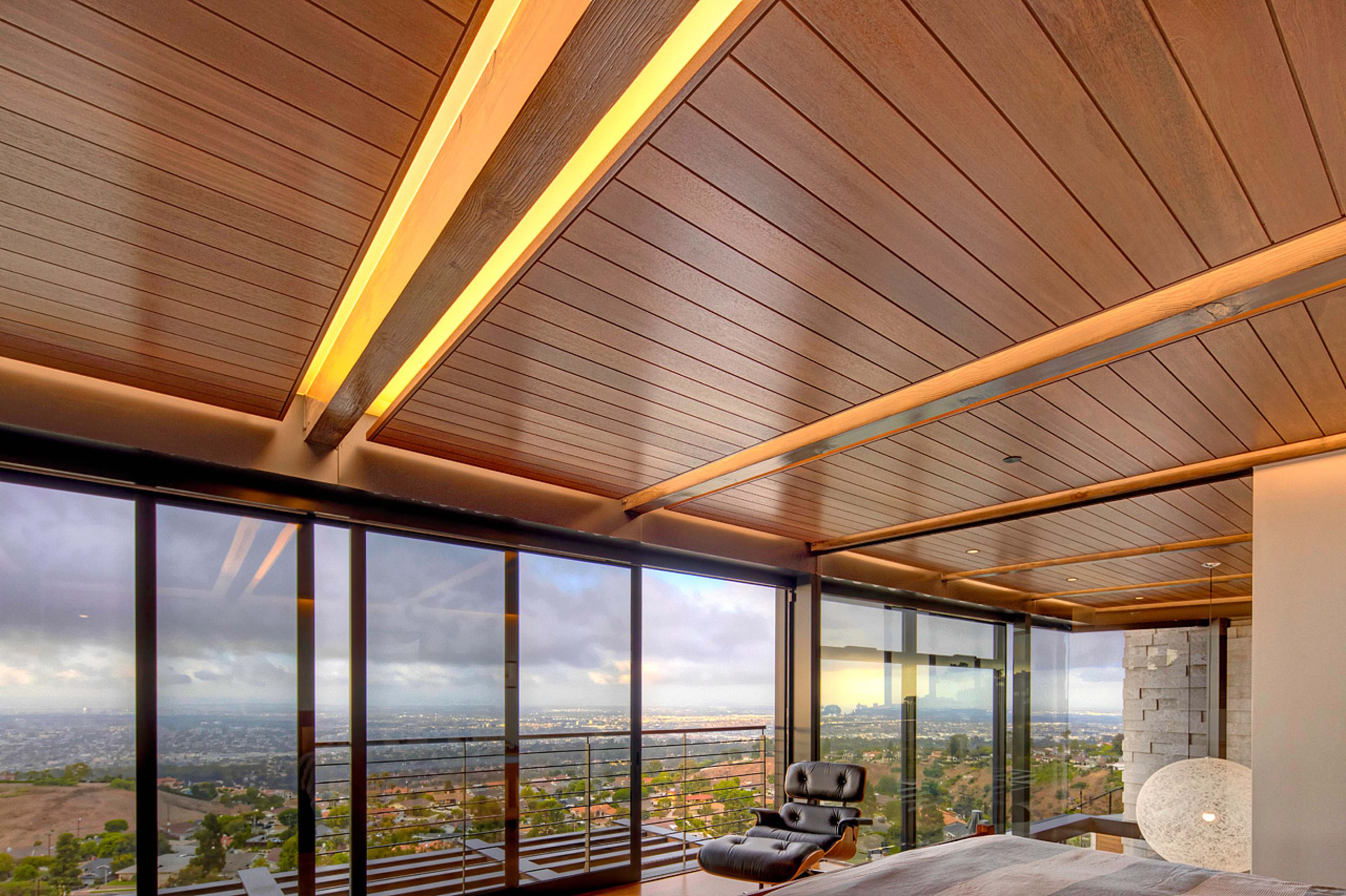 A modern bedroom with a wooden ceiling, large glass windows overlooking a city landscape, and a stylish black chair in the corner.