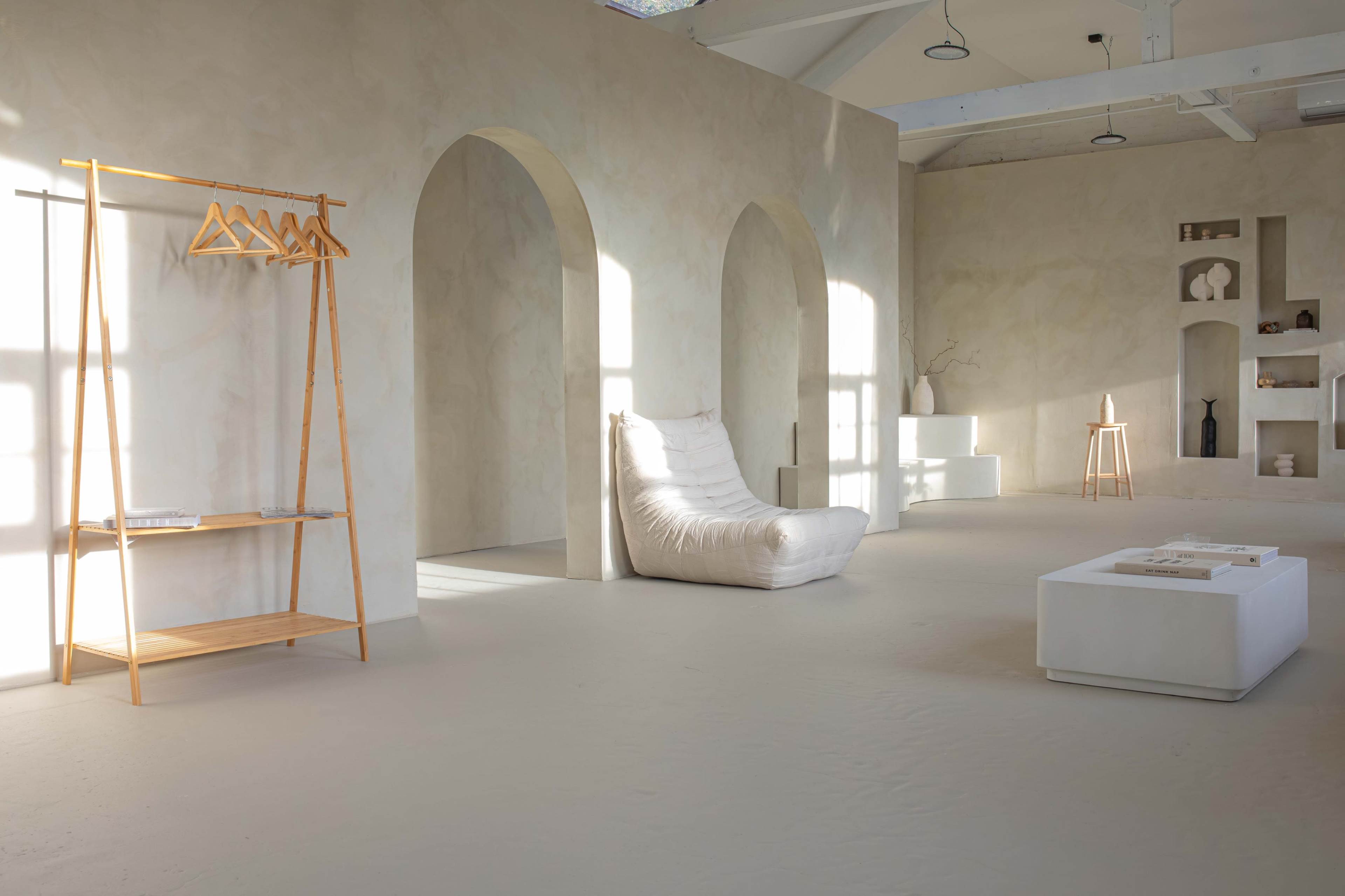 The image shows a spacious, minimalist interior featuring a white lounge chair, a wooden coat rack, and shelves displaying decorative objects against a light-colored wall.