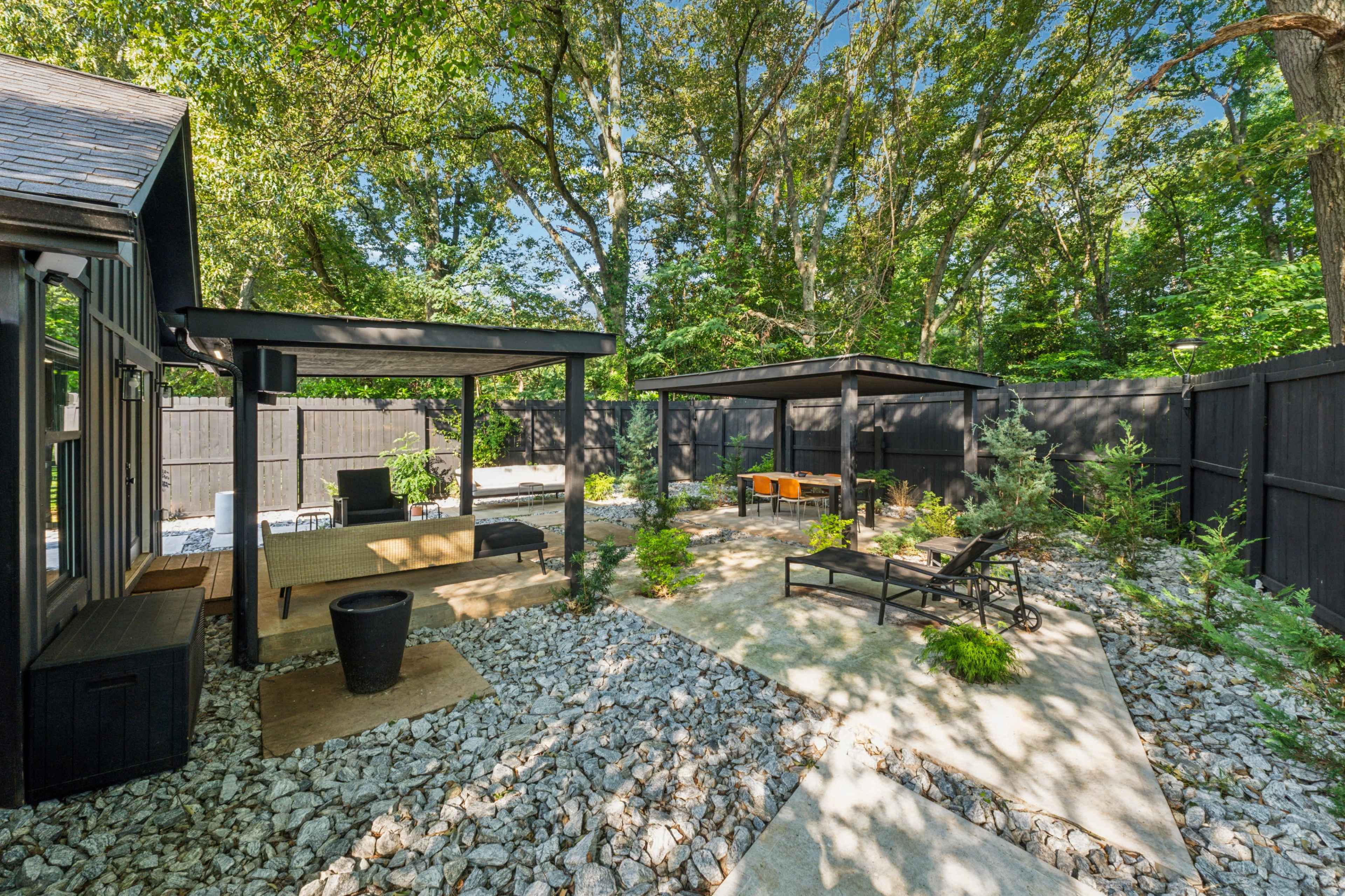 The image depicts a landscaped outdoor space featuring two shaded structures, gravel pathways, and a mix of greenery on a sunny day.