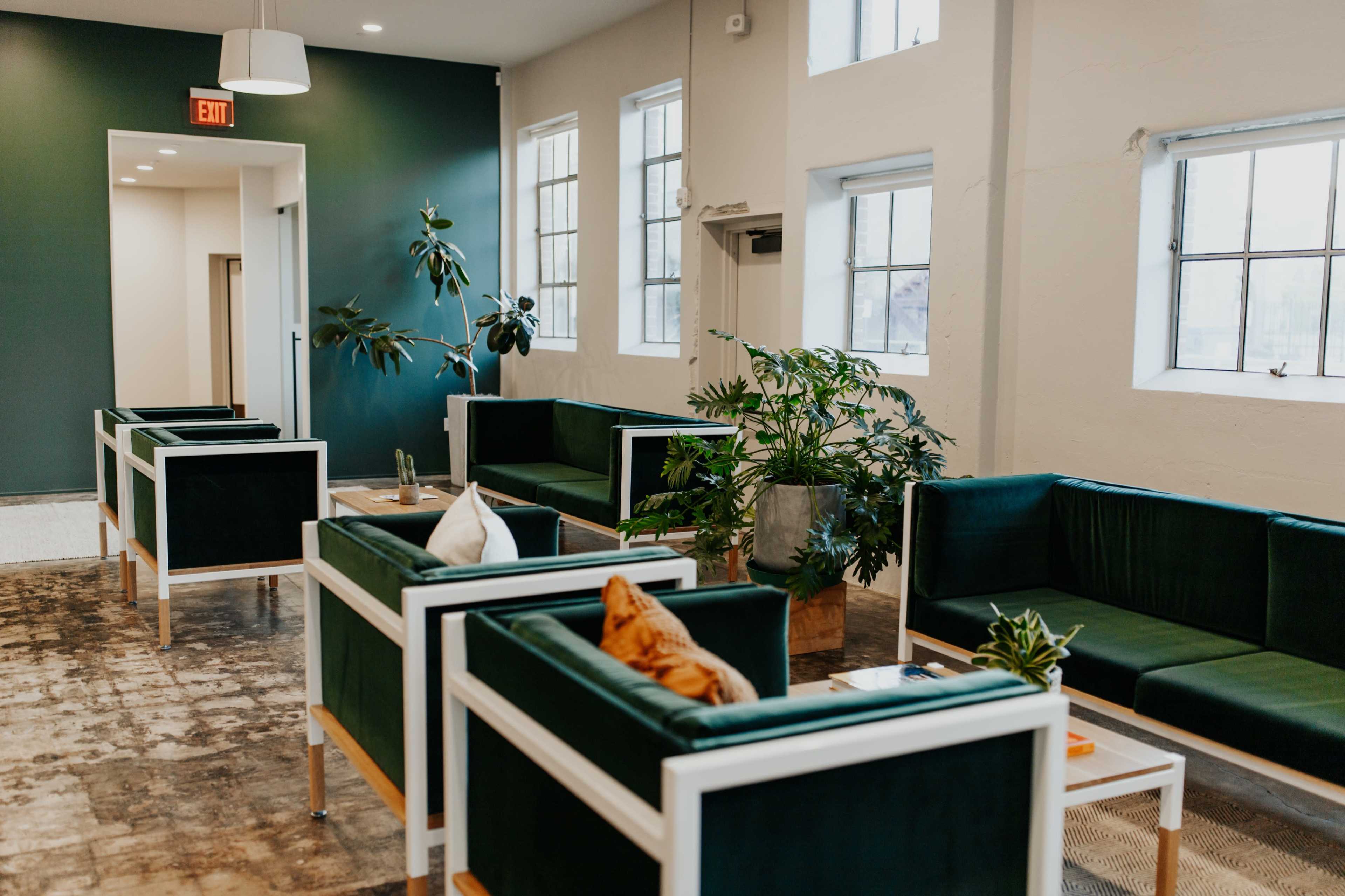 A modern lounge area with green couches arranged around potted plants and large windows.