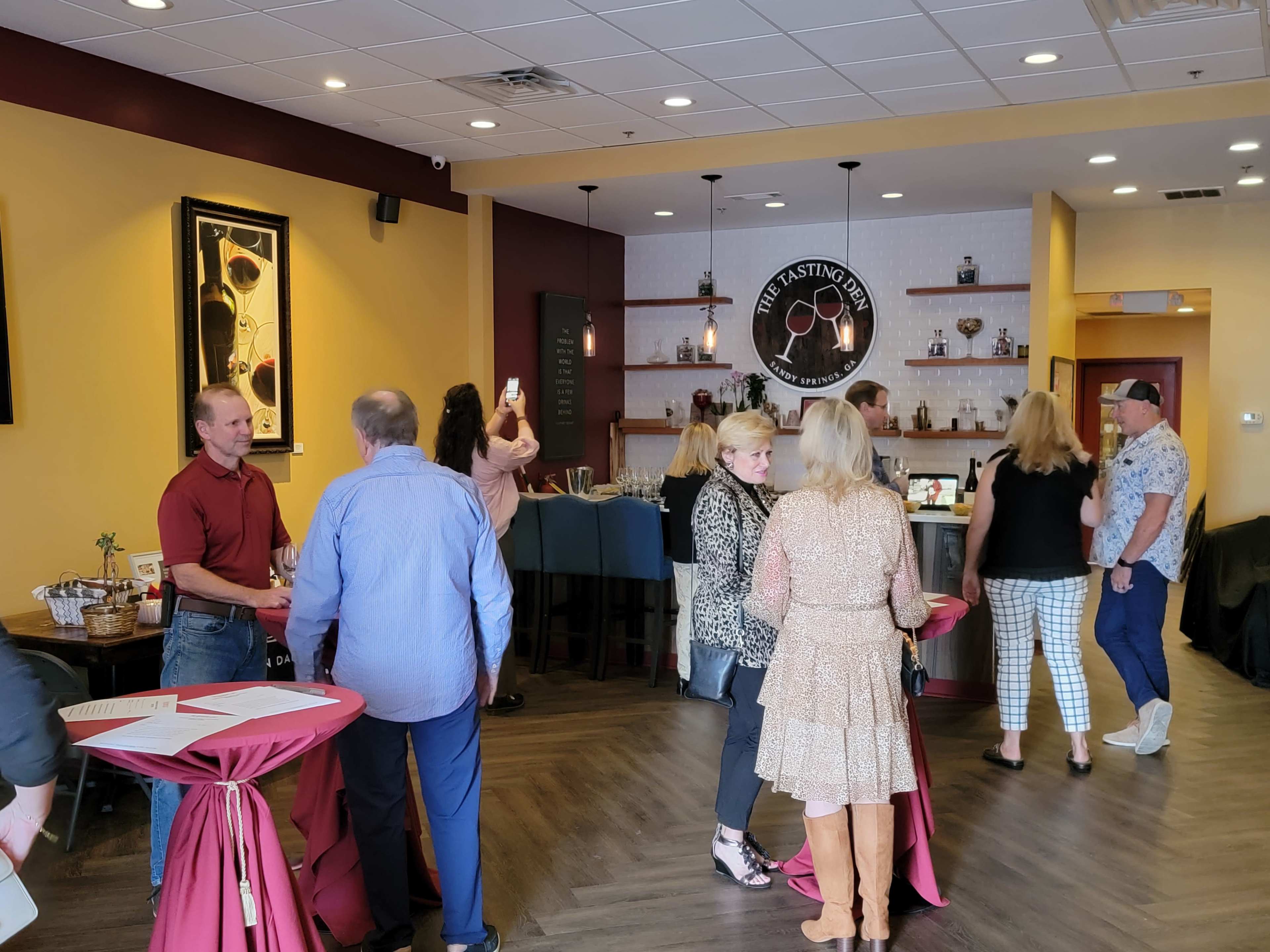A group of people socialize in a wine tasting room, with some standing and others at tables, while a bar area with various bottles is visible in the background.
