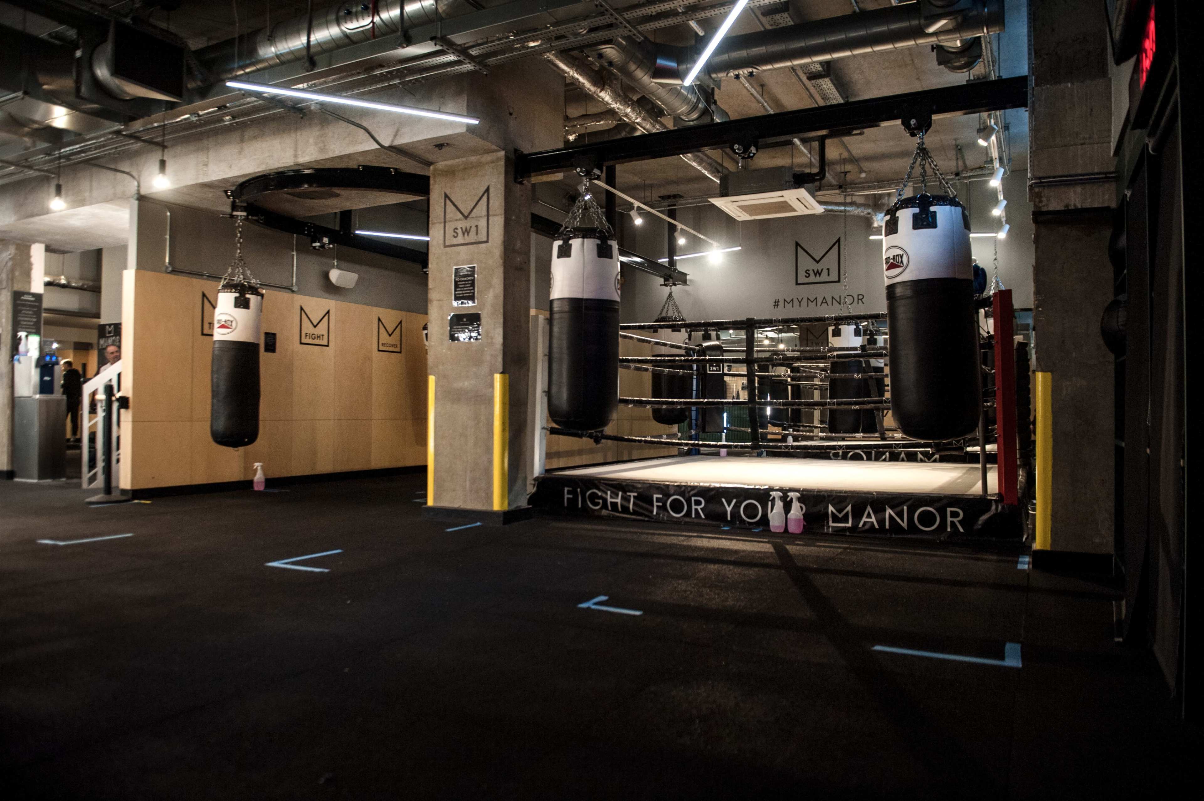 The image shows an indoor boxing gym featuring several hanging punching bags and a boxing ring in the background.