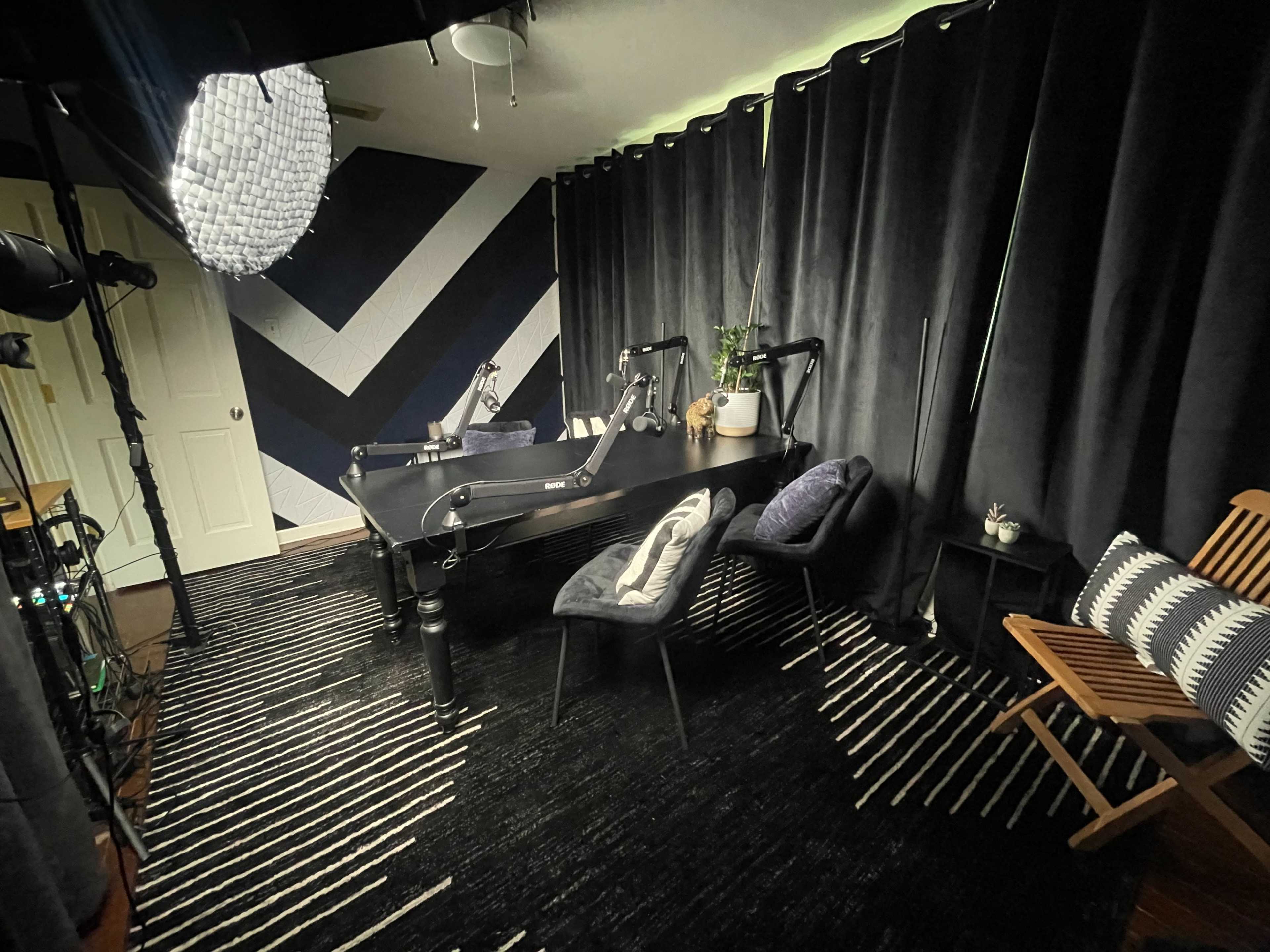 The image shows a modern podcasting or recording studio featuring two chairs, a table, and dark curtains against a chevron-patterned wall.