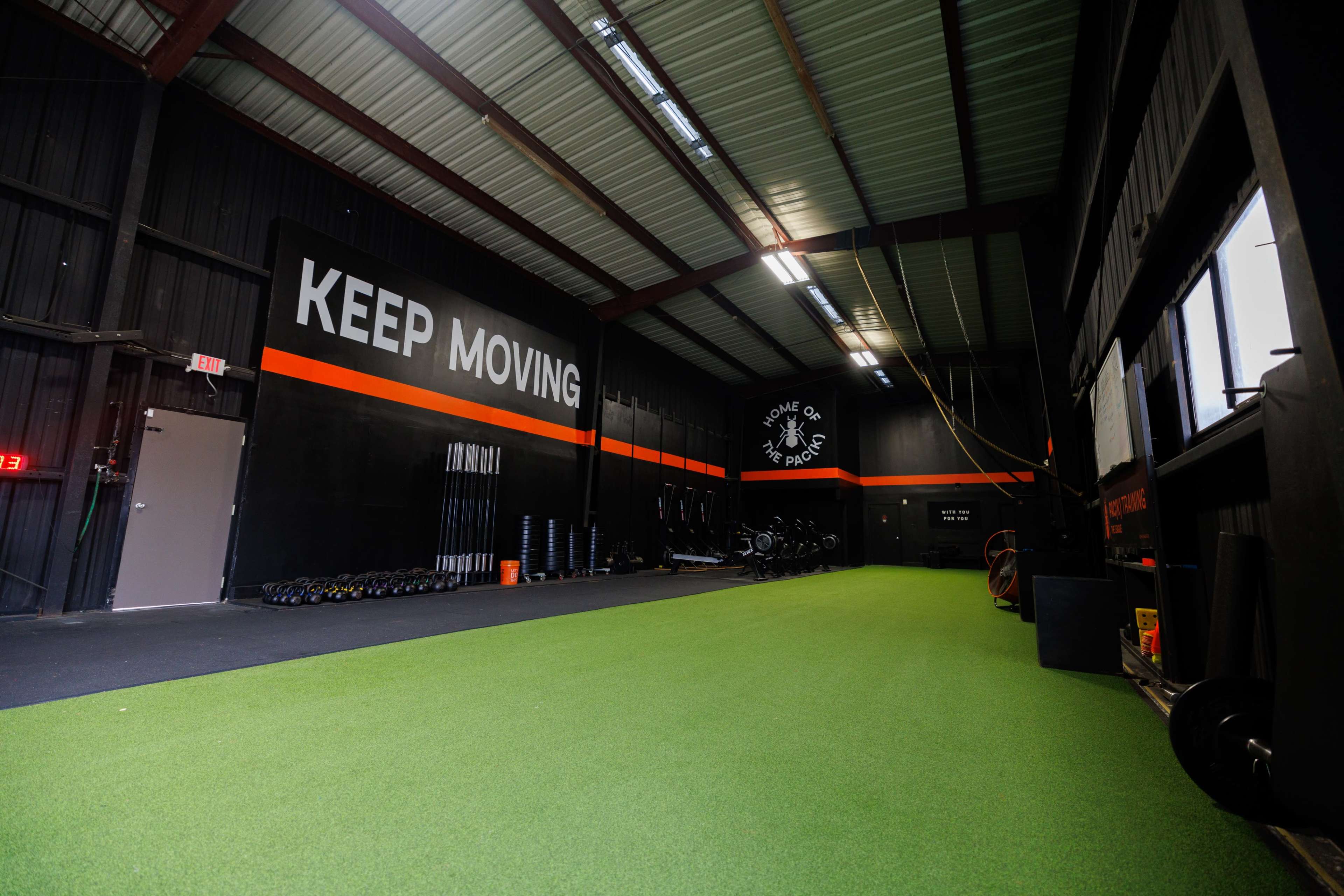 A spacious gym features a green turf floor, black walls with motivational text, and equipment arranged along the sides.