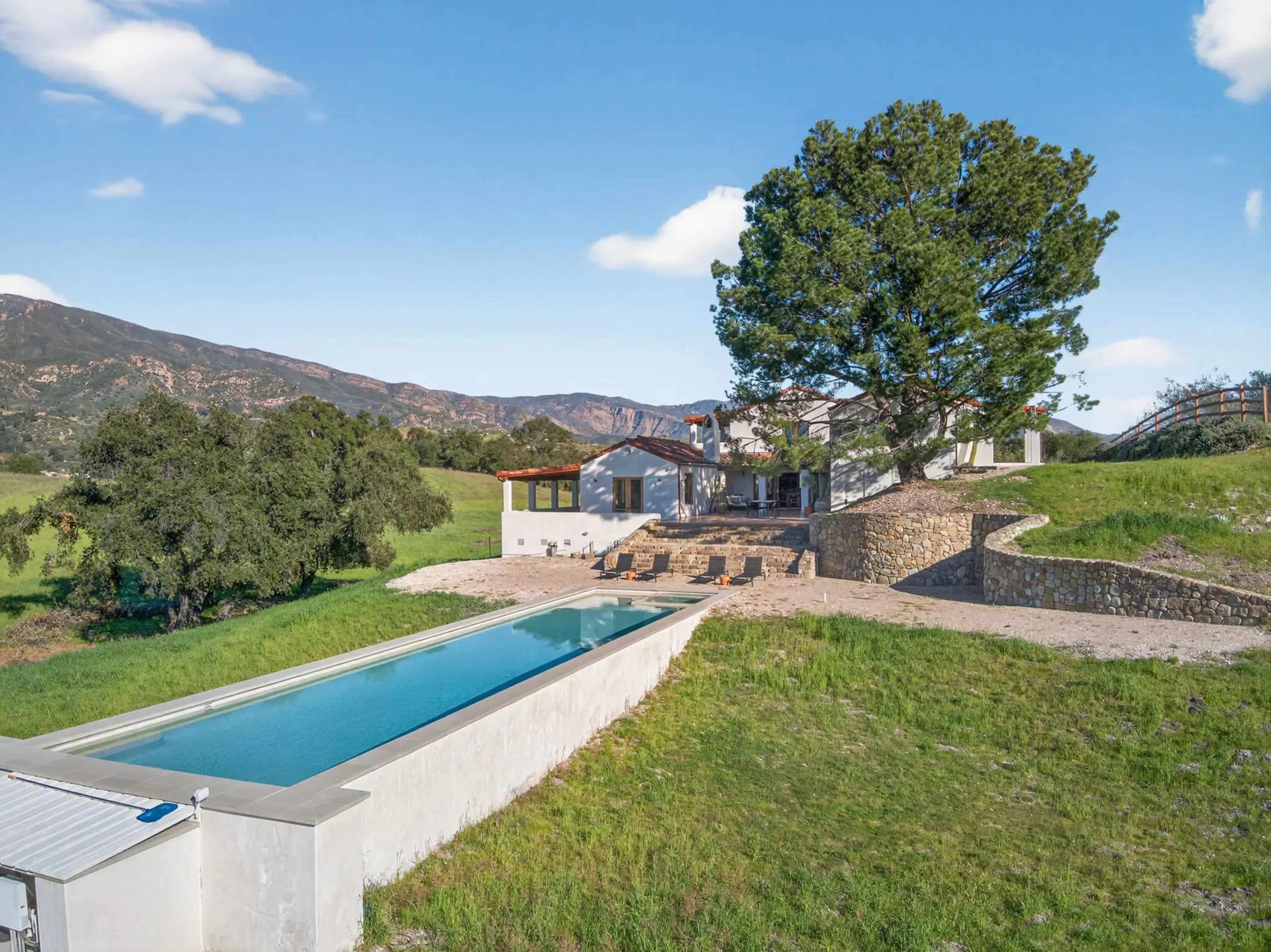 A modern house with a long, narrow swimming pool is situated in a green landscape with mountains in the background.