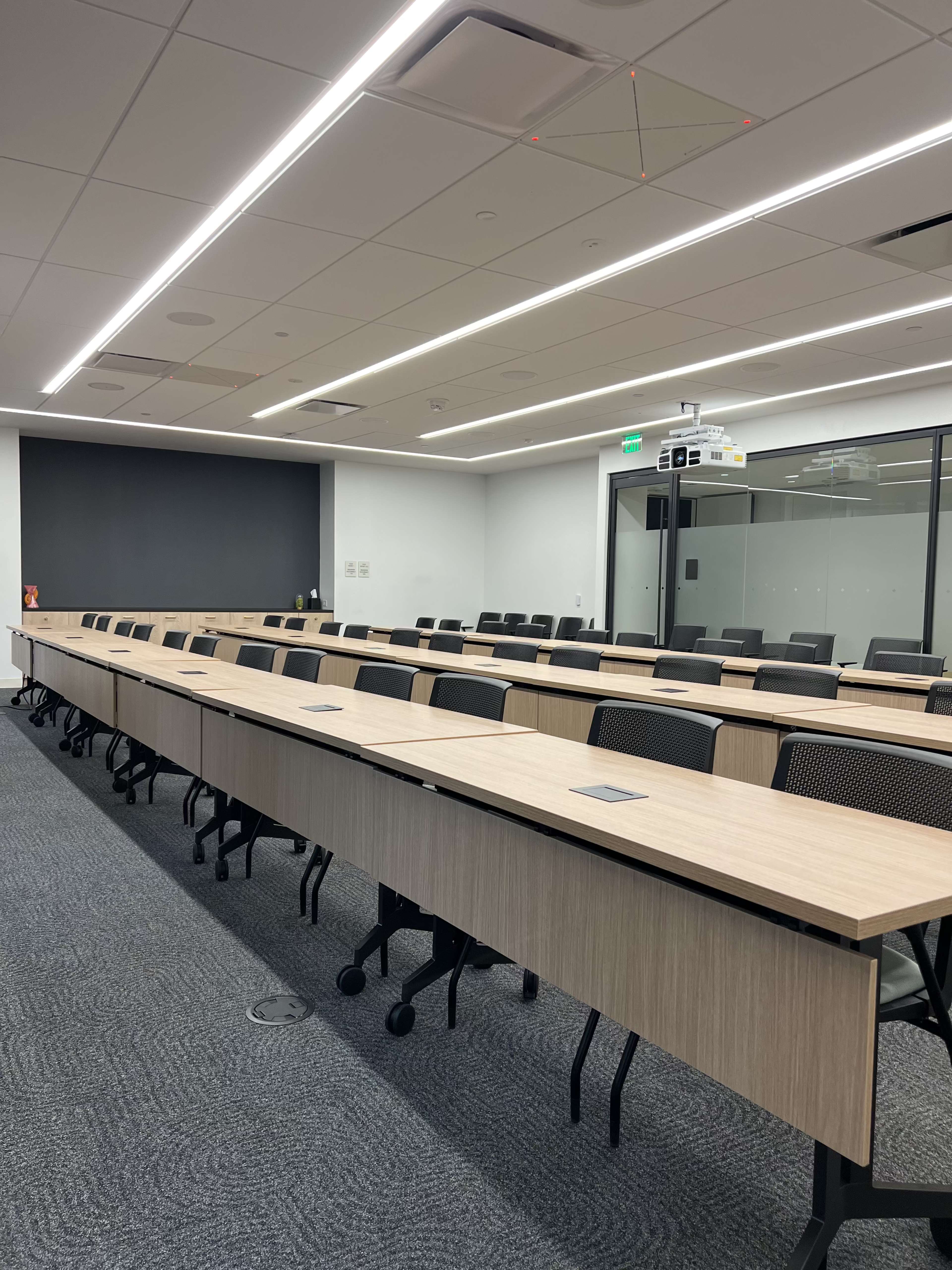 The image shows a modern conference room with multiple rows of long tables and chairs, featuring overhead lighting and a wall-mounted projector.