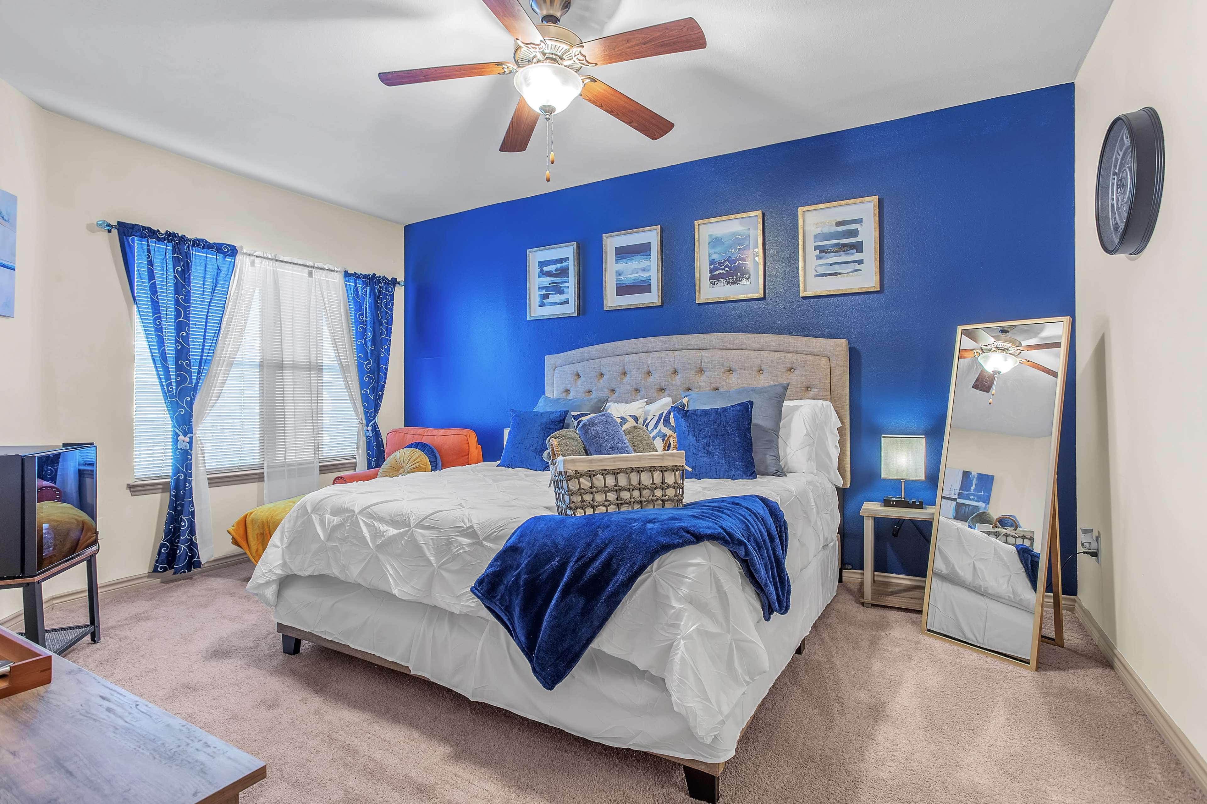 The image shows a bedroom featuring a large bed with a tufted headboard, blue walls, colorful accents, and a ceiling fan.
