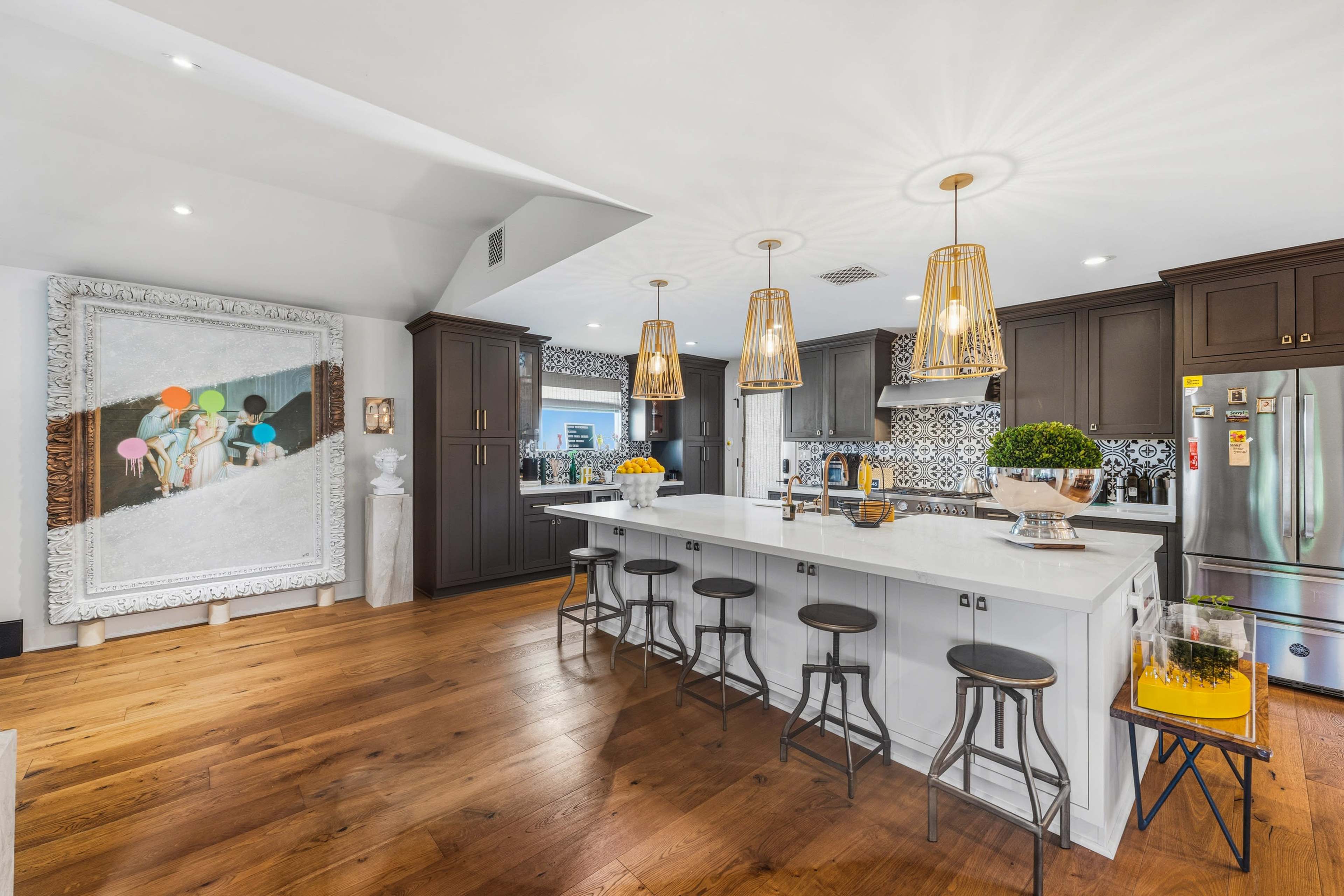 The kitchen features a large island with bar stools, modern cabinetry, and a decorative painting on the wall, all illuminated by stylish pendant lights.