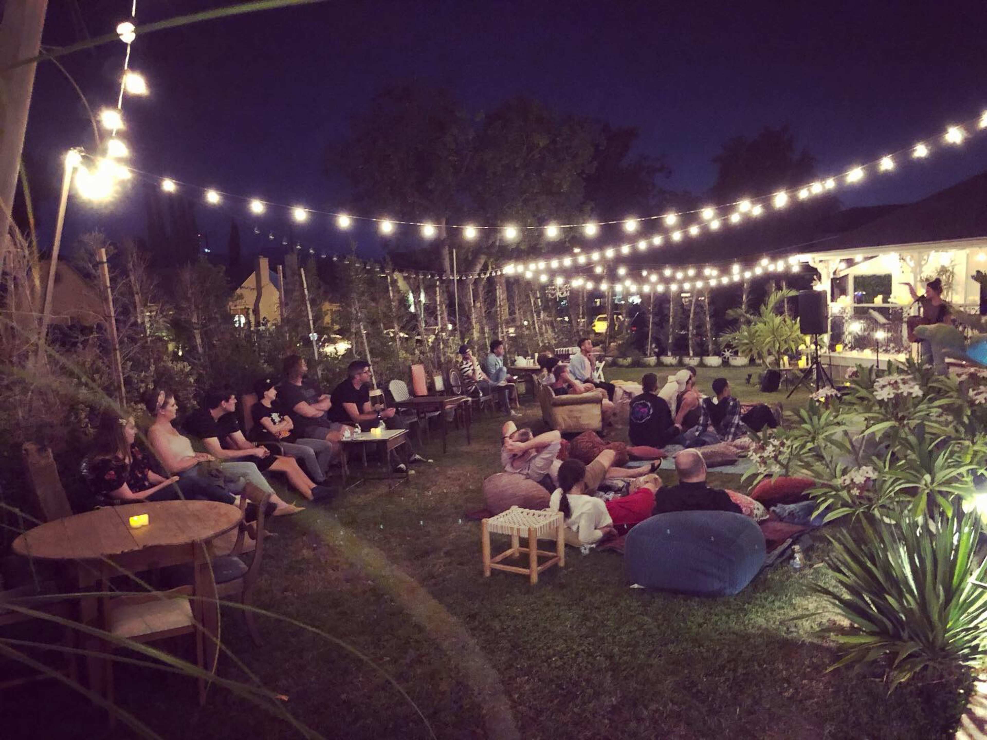 A group of people sits in a garden under string lights, gathered for an outdoor performance.