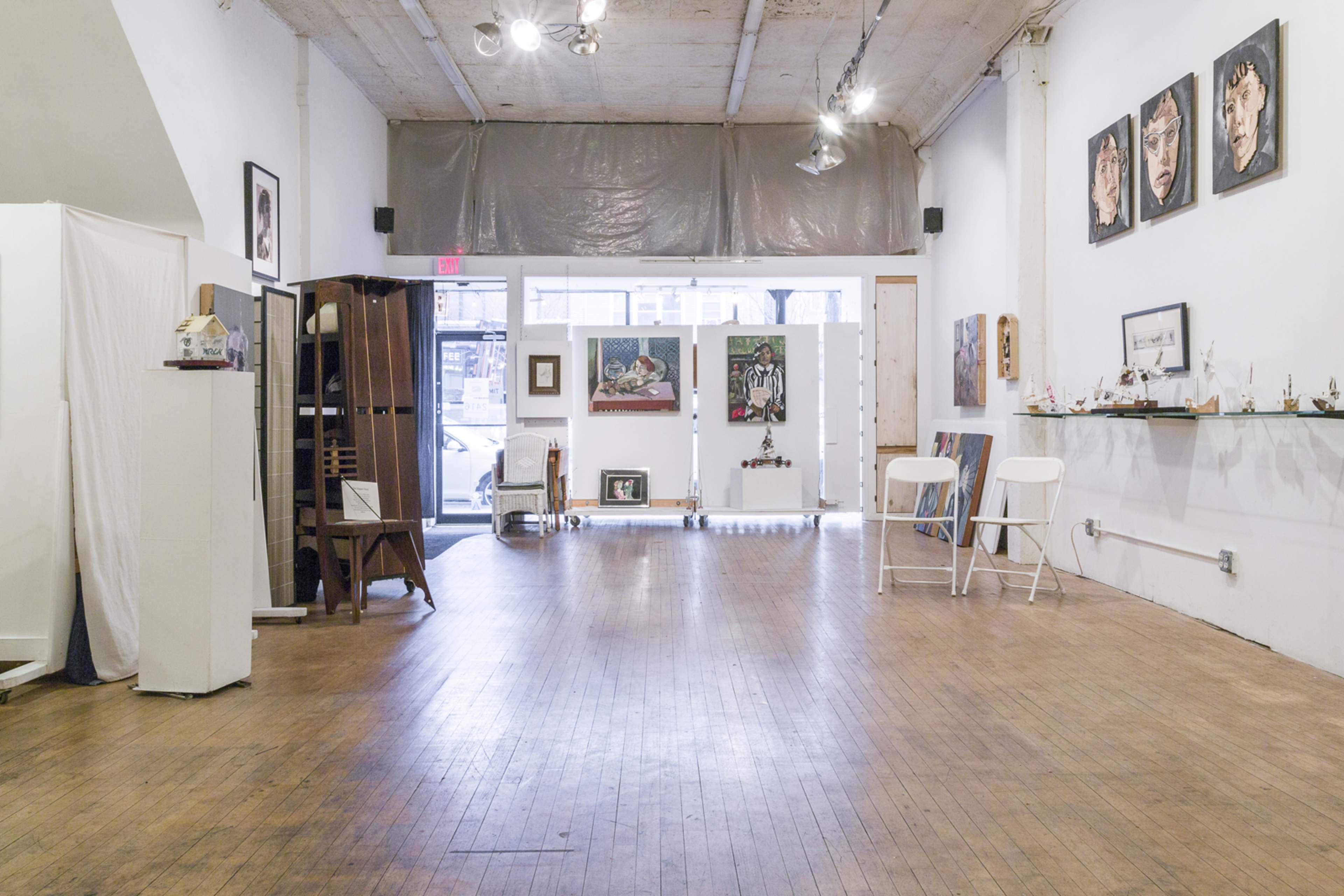 The image shows a spacious, well-lit art gallery with wooden floors and various artworks displayed on the walls and shelves.