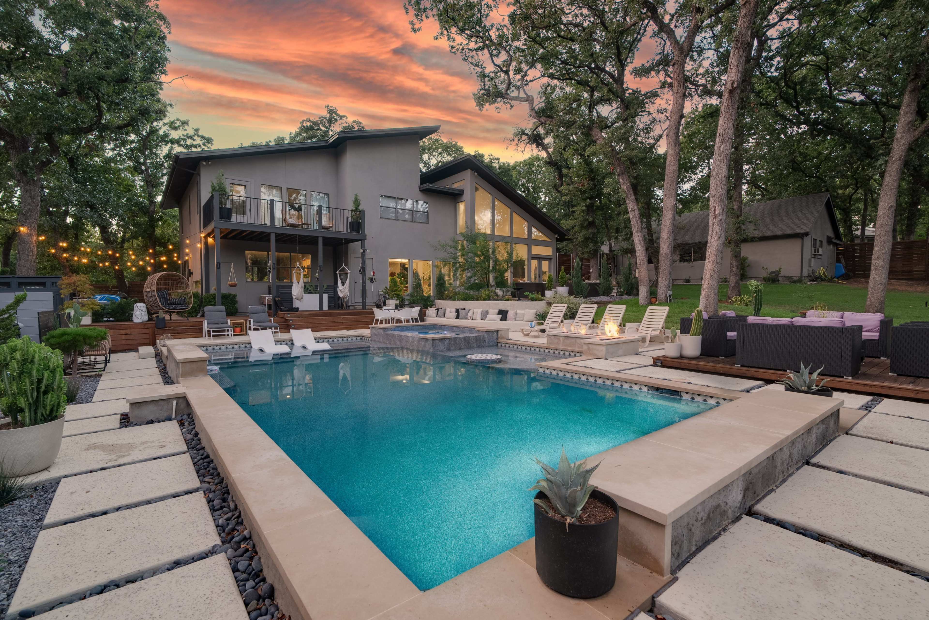 The image shows a modern house with a large swimming pool surrounded by lounge chairs and landscaped greenery at sunset.