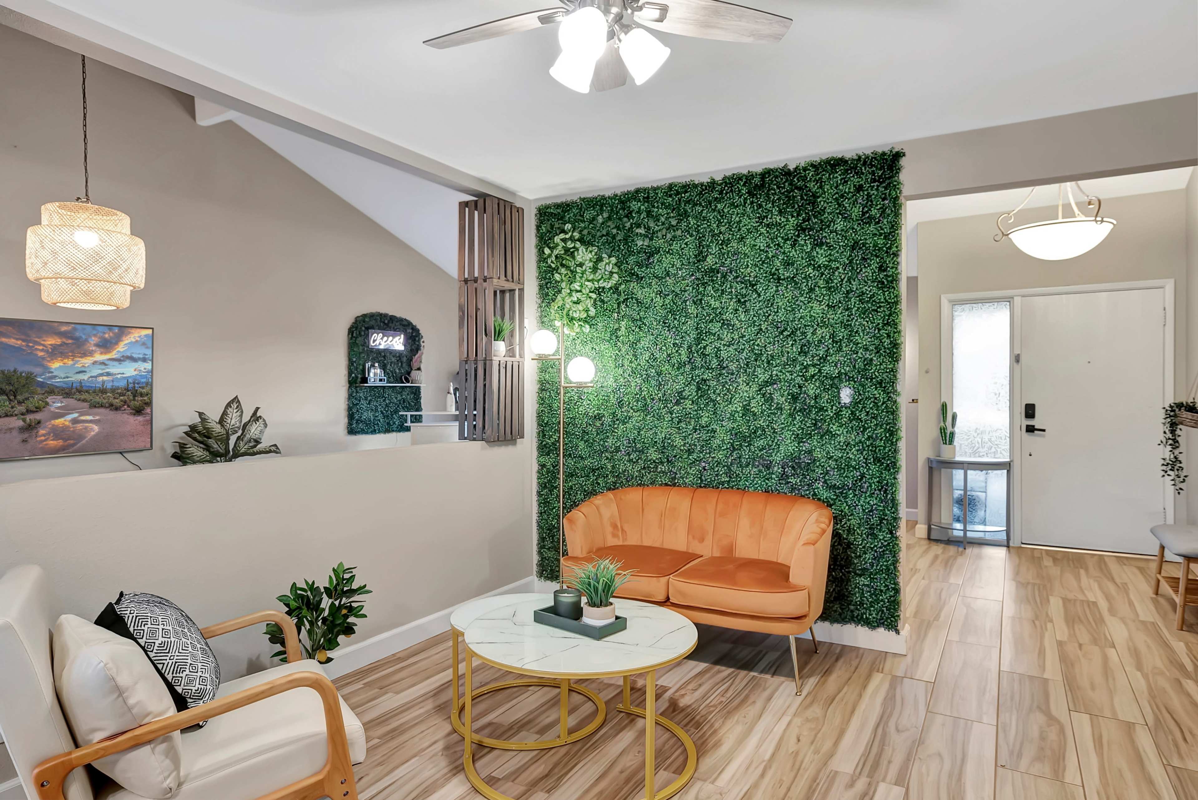 A cozy living space features a green wall plant backdrop, an orange couch, a round coffee table, and a light fixture above.