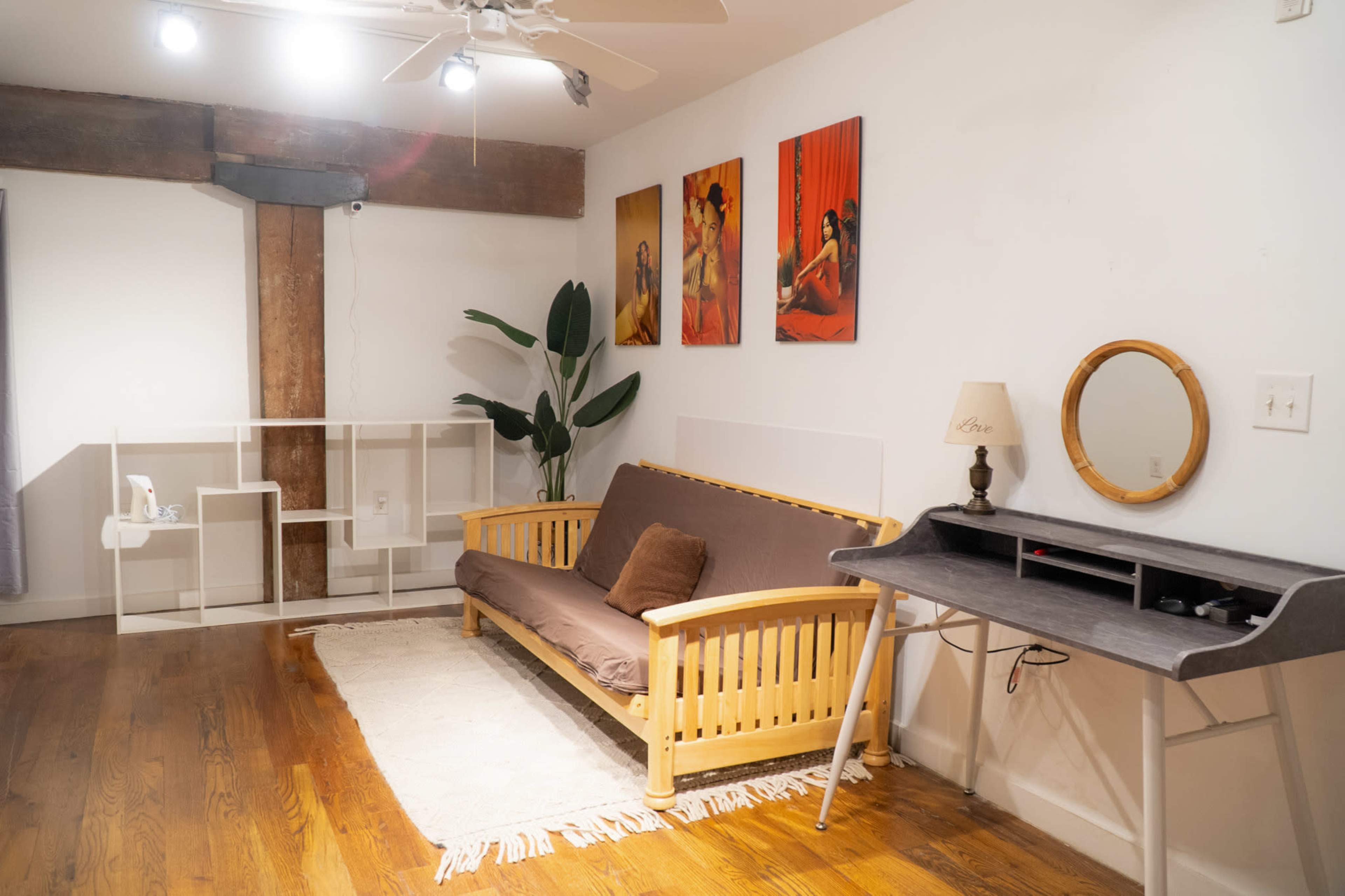 The image shows a simple living space featuring a wooden futon, a desk with a round mirror, and three colorful wall art pieces.