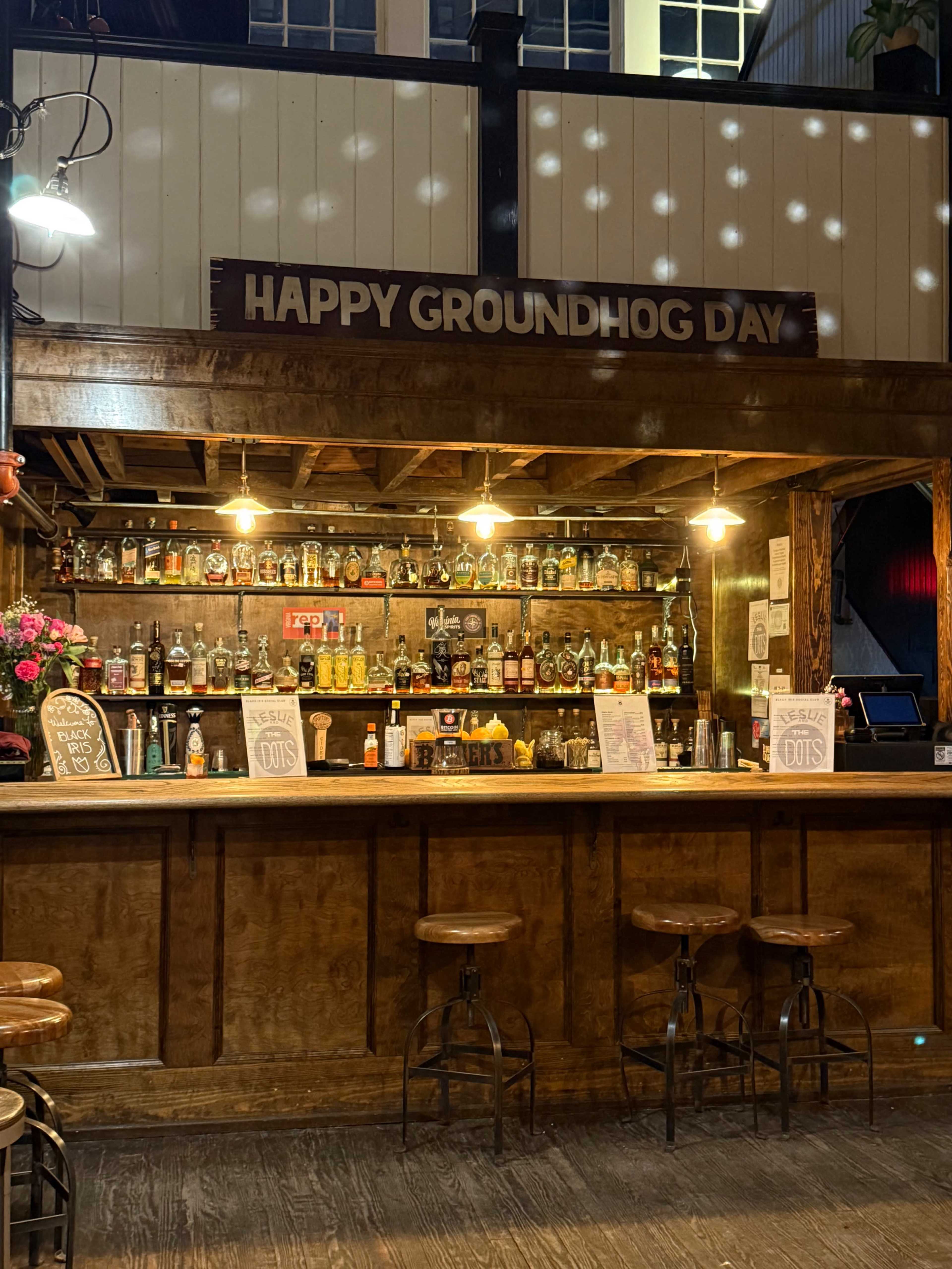 The image shows a wooden bar with a sign reading "Happy Groundhog Day" above shelves lined with various bottles of liquor, and empty bar stools in front.