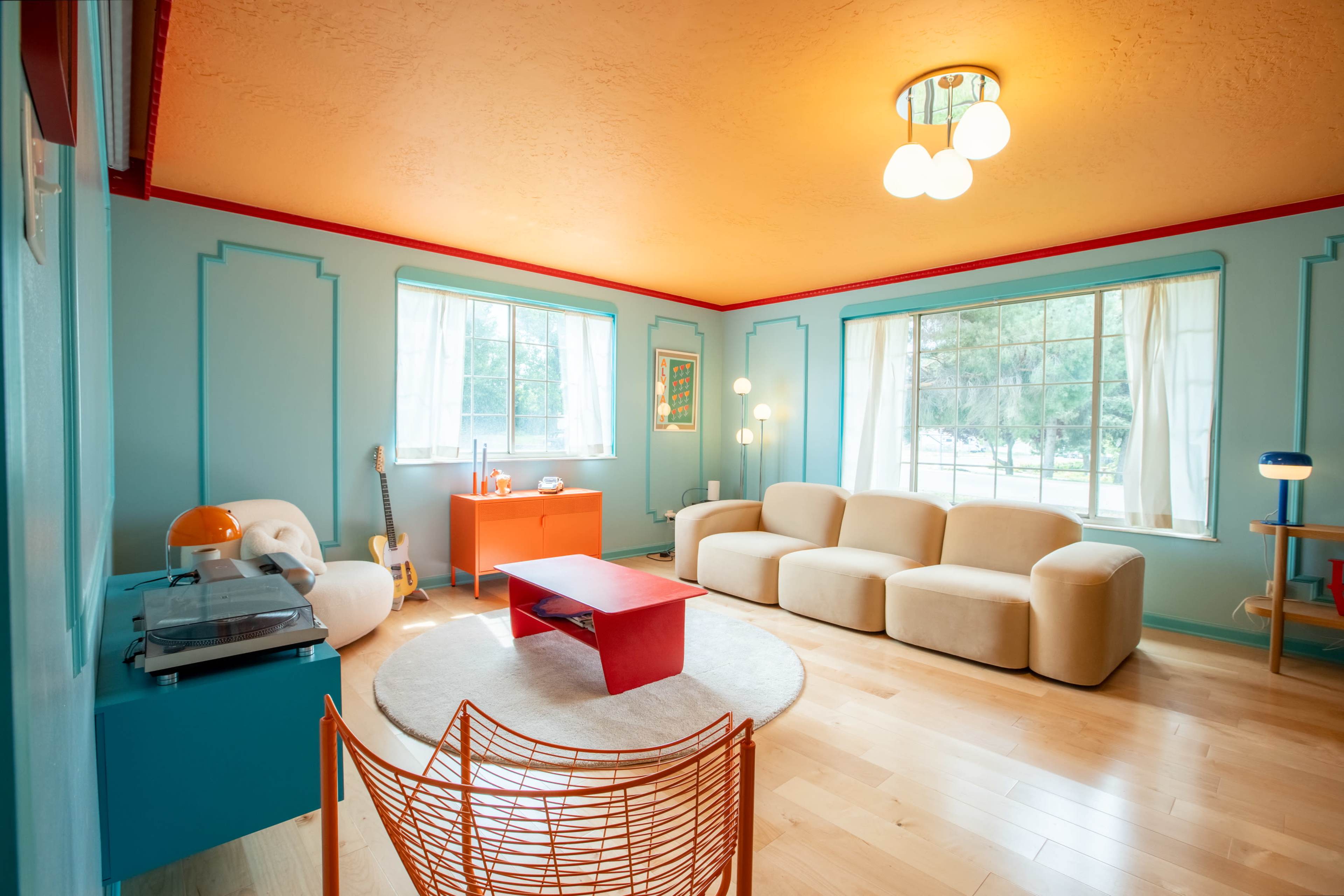 The room features a modern design with an orange ceiling, turquoise walls, and a mix of furniture including a beige sofa, a red coffee table, and a decorative wire chair.