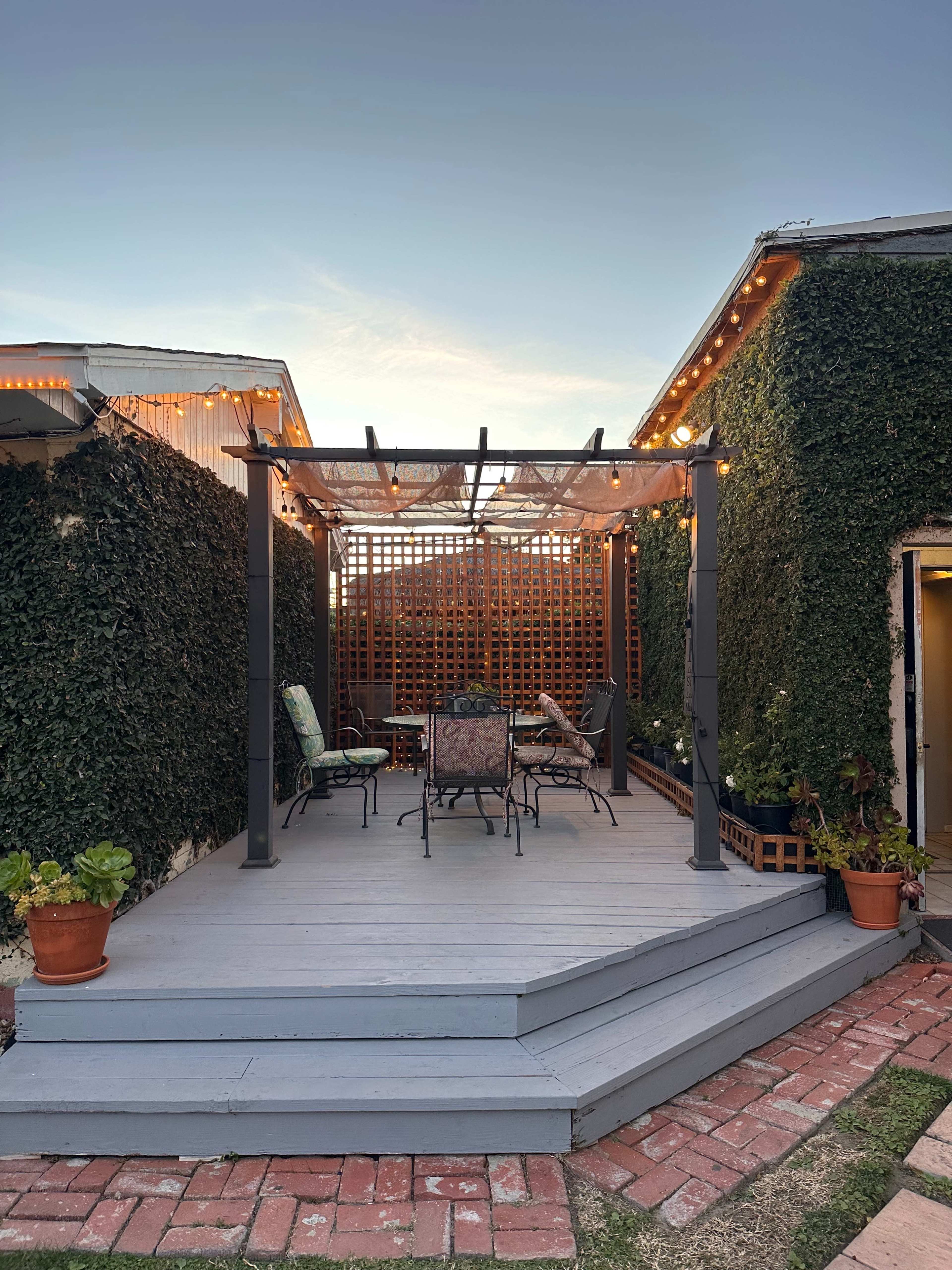 A cozy patio area with a wooden pergola, surrounded by green ivy, features a table and chairs under warm string lights.