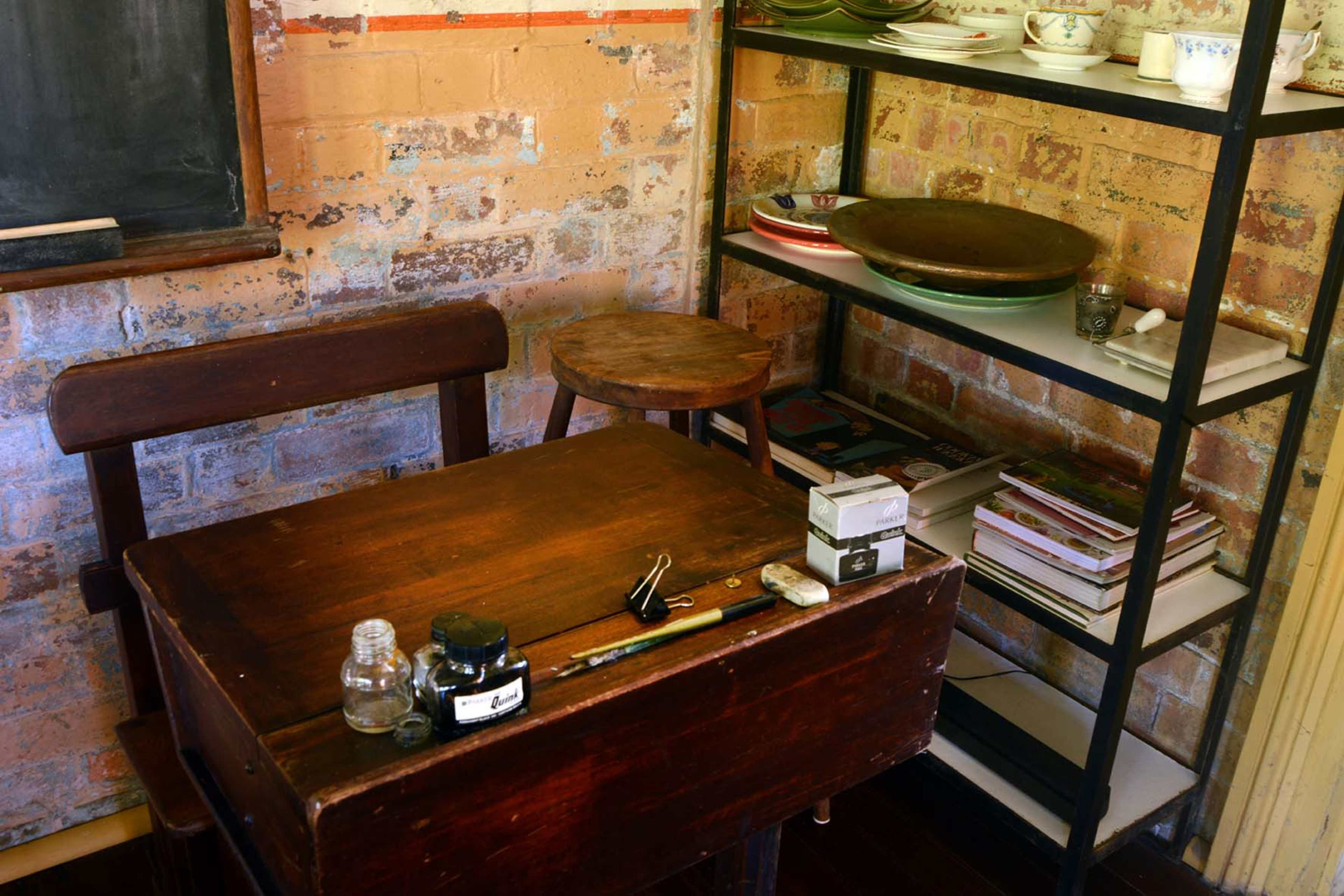 A wooden desk with various writing tools and ink sits next to a two-tiered shelf displaying plates and books in a room with exposed brick walls.