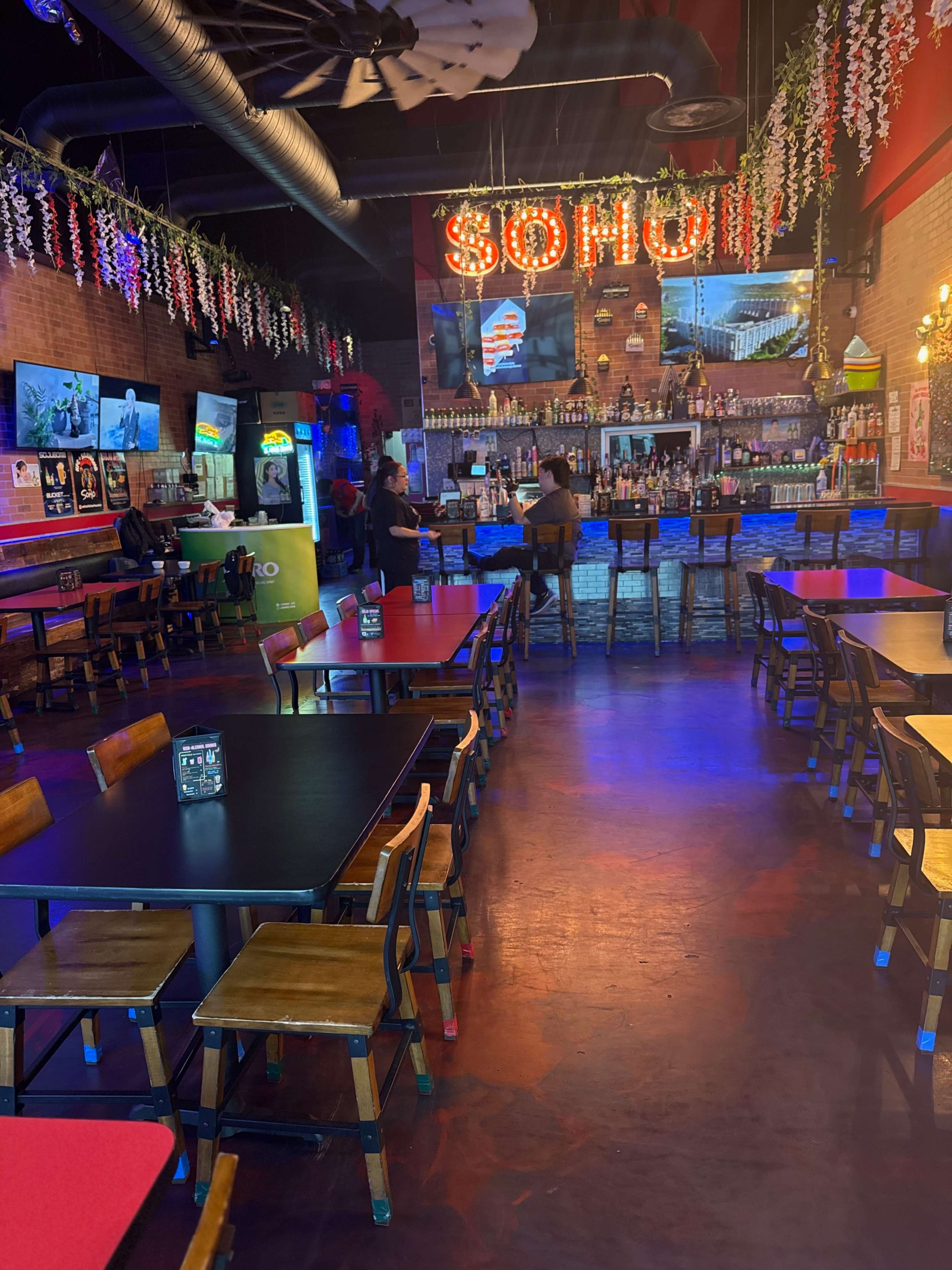 The interior of a bar-restaurant is shown with tables and chairs arranged in a mostly empty space, a brightly lit bar area in the background, and vibrant décor on the walls.