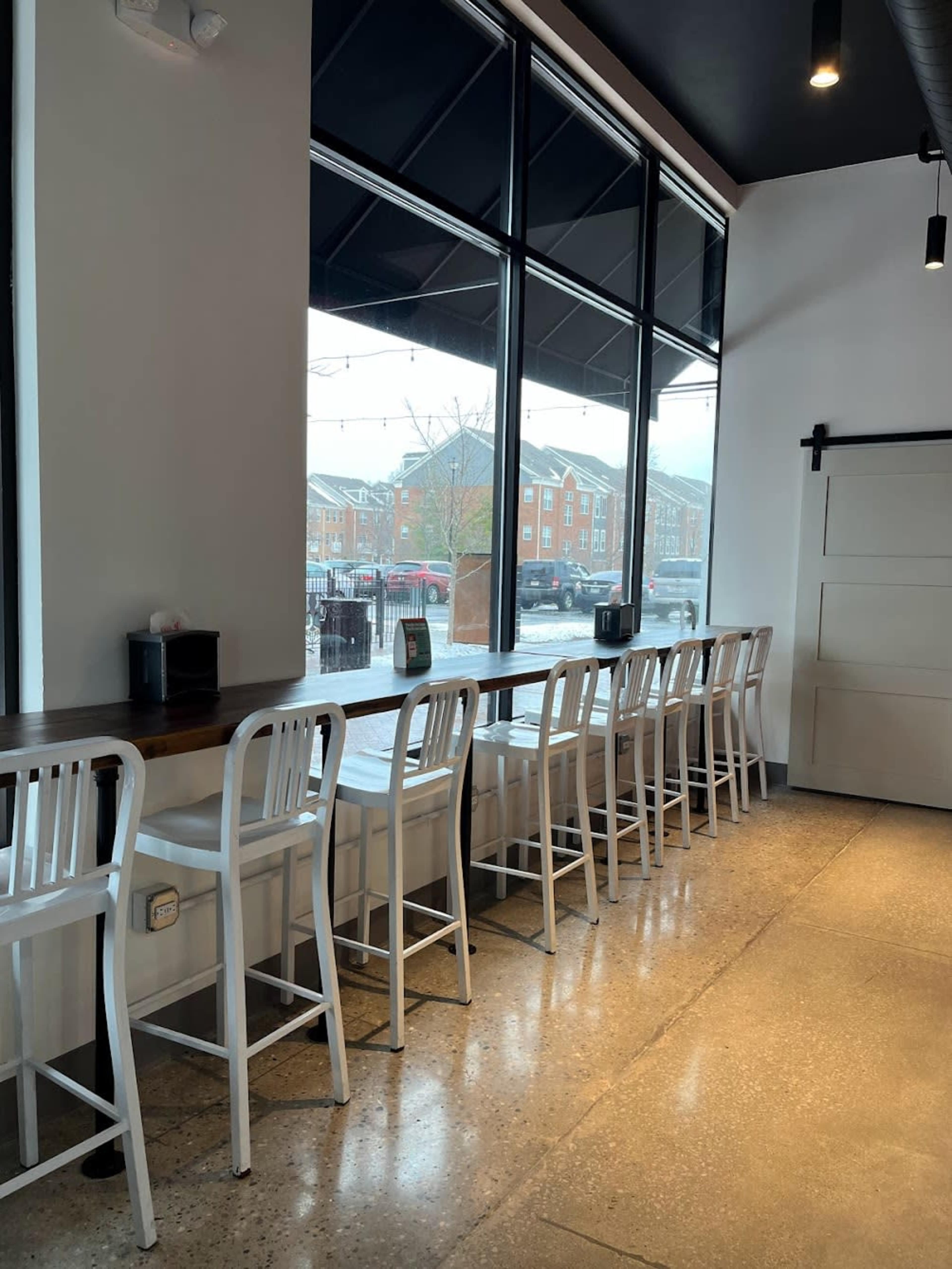 A row of white metal bar stools is lined up against a large window in a café, overlooking a street with brick buildings in the background.