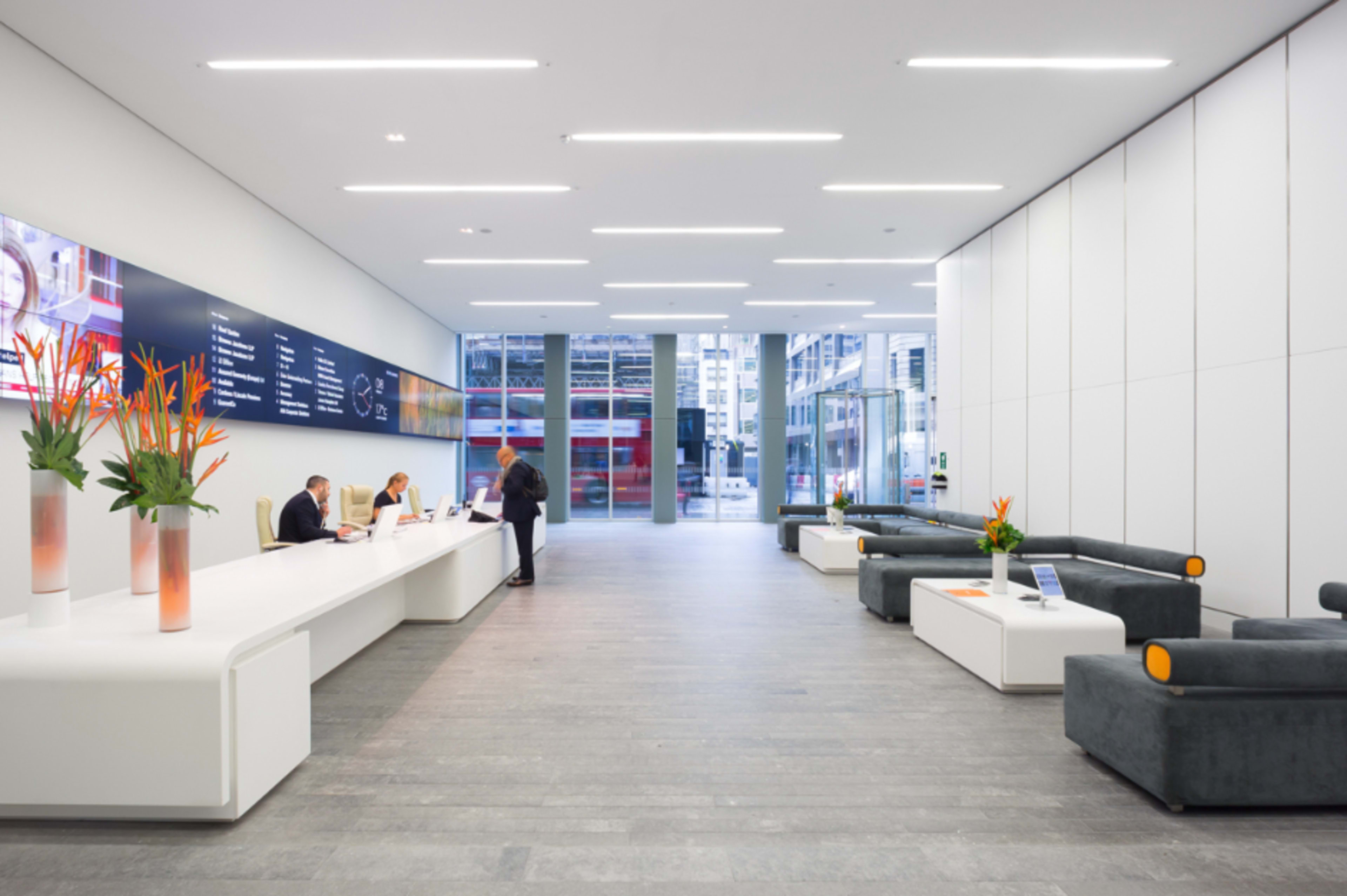 The image features a modern reception area with a sleek white desk, two individuals working on laptops, floral arrangements, and gray seating against a minimalist backdrop of large windows.