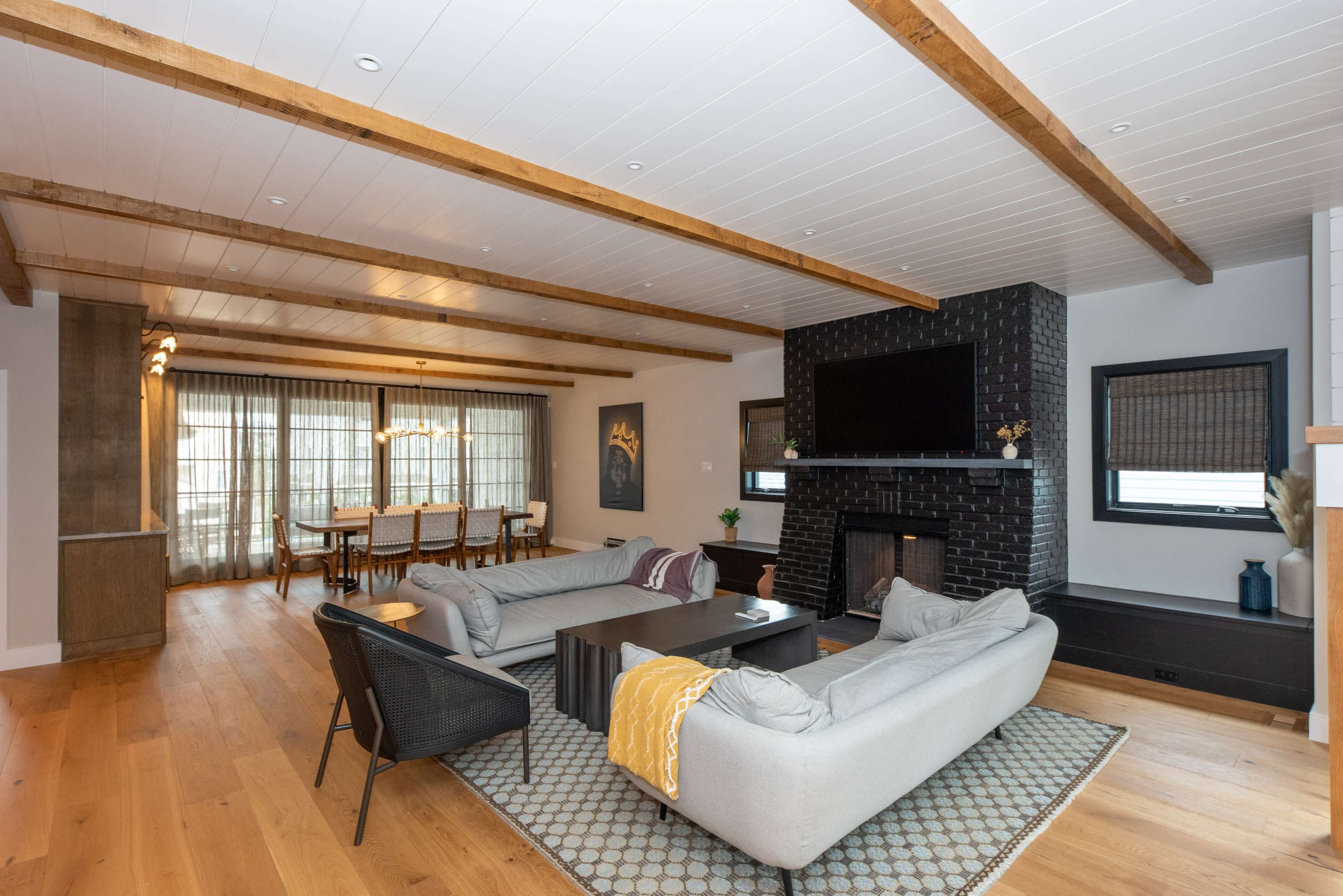 A spacious living room features a gray sectional sofa, a dark stone fireplace, and large windows with sheer curtains, leading to a dining area with a long wooden table.