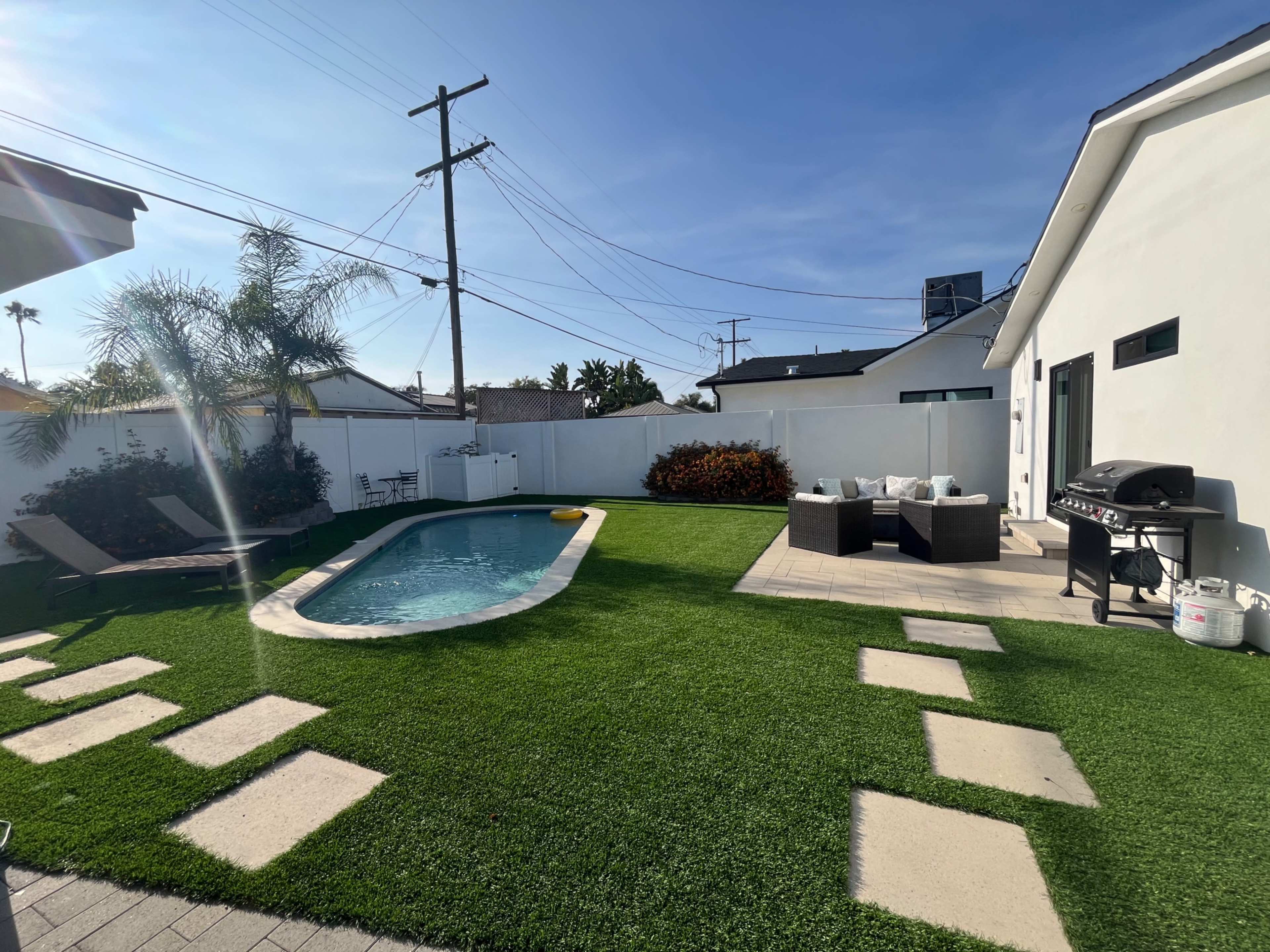 A backyard with a swimming pool, a patio area with a grill, and artificial grass bordered by stone pathways and shrubs.