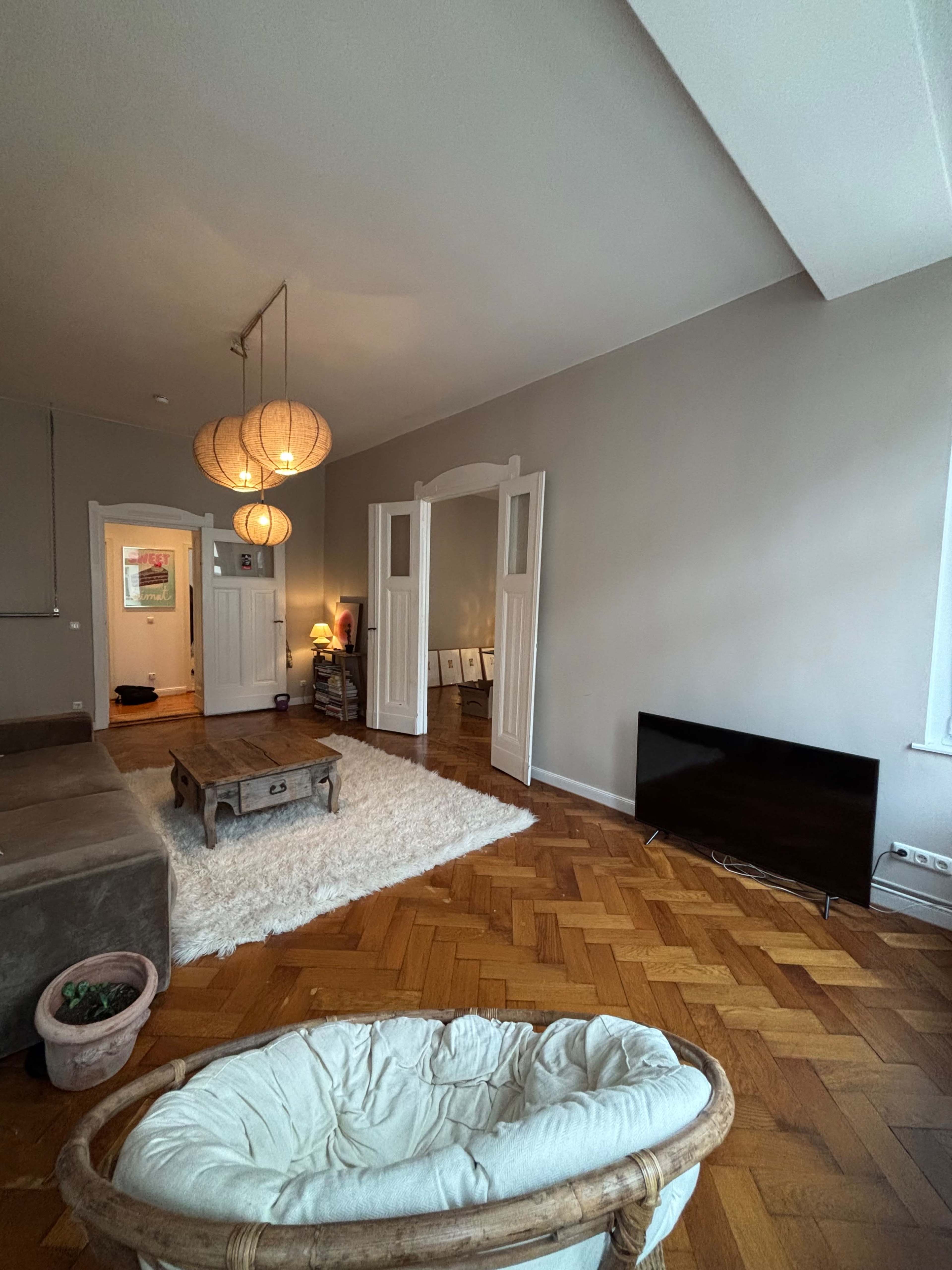 The image shows a spacious living room with a light gray wall, wooden flooring, a plush rug, a couch, a coffee table, and a television against one wall.