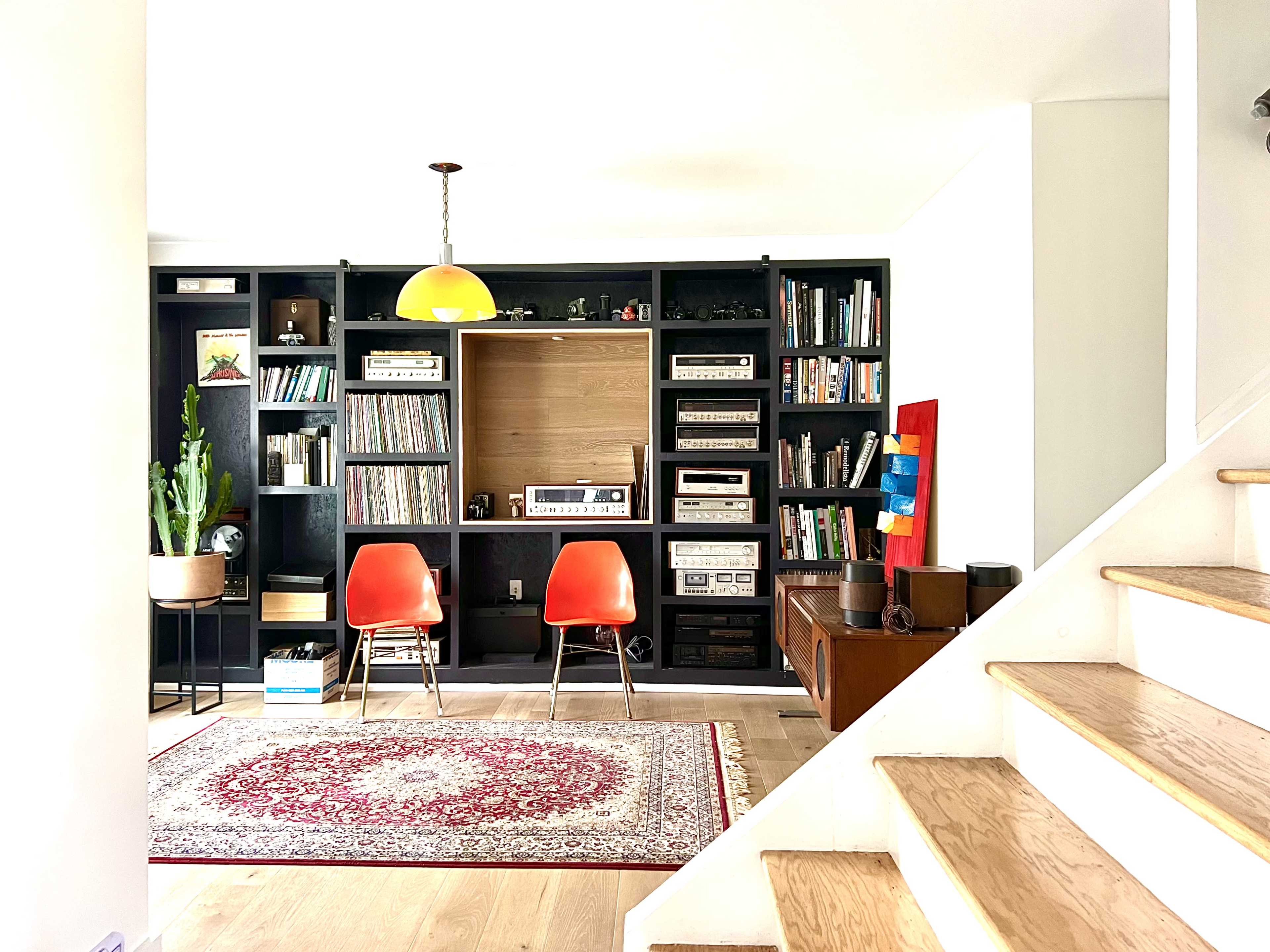 A cozy living area featuring a record player and vinyl collection, two orange chairs, a bookshelf filled with books, and a staircase leading to another level.
