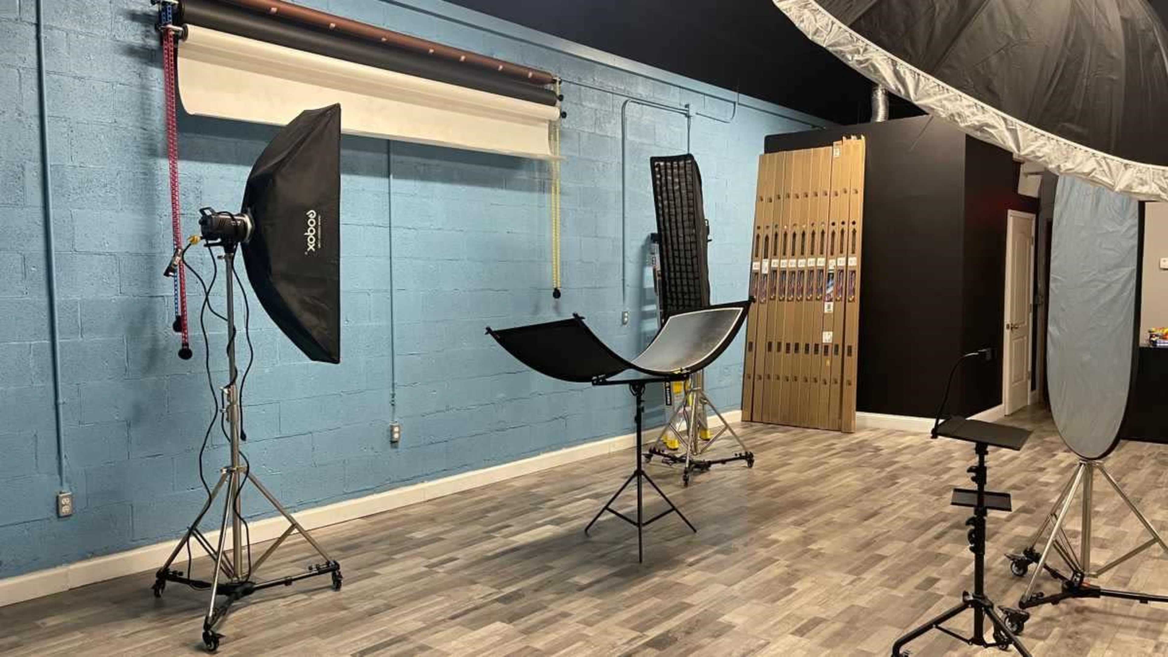 The image shows a photography studio with various equipment, including light modifiers and supports, set against a blue wall.