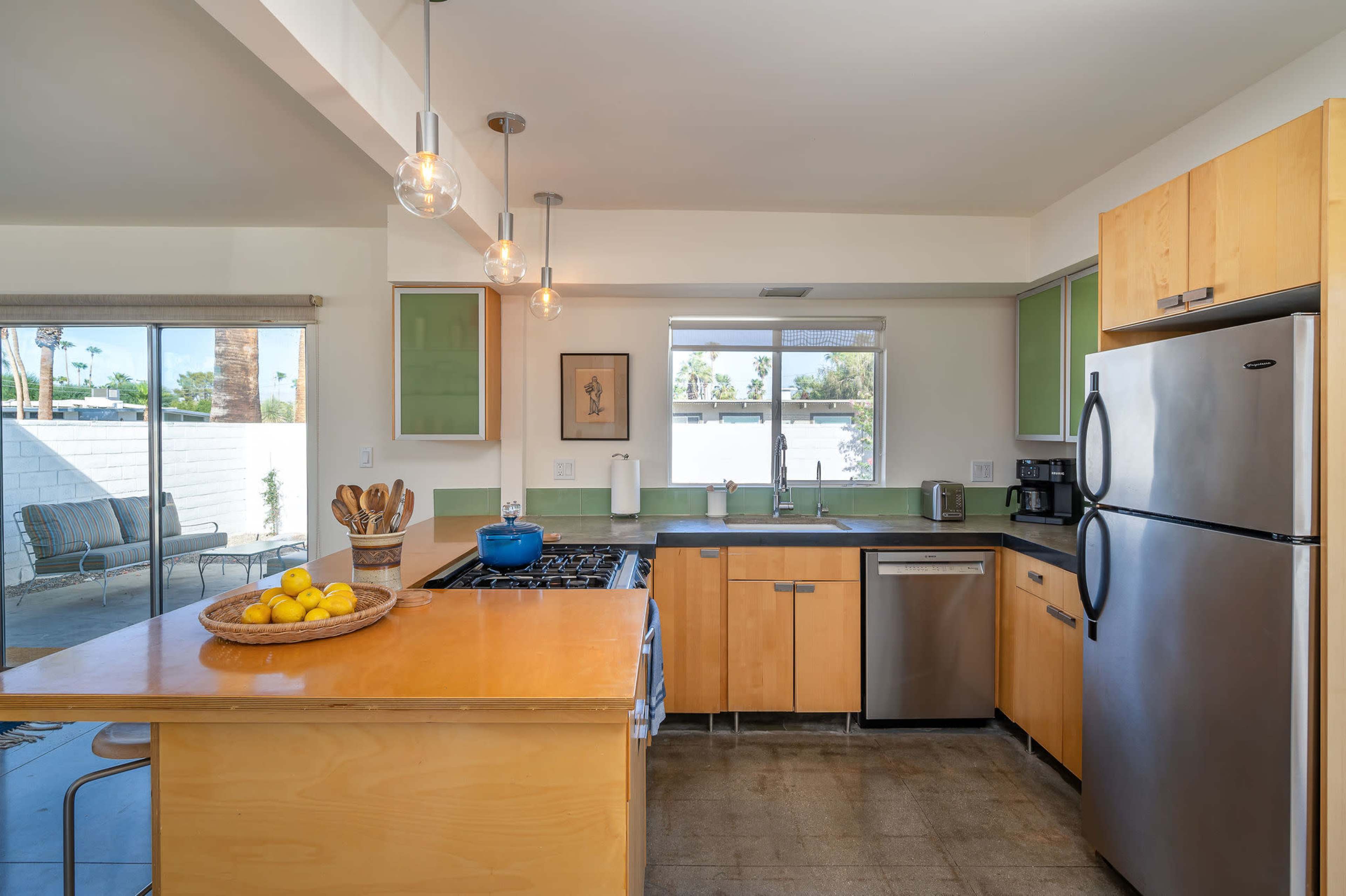 A modern kitchen features wooden cabinetry, a stainless steel refrigerator, and a large island with a fruit bowl, illuminated by pendant lights.