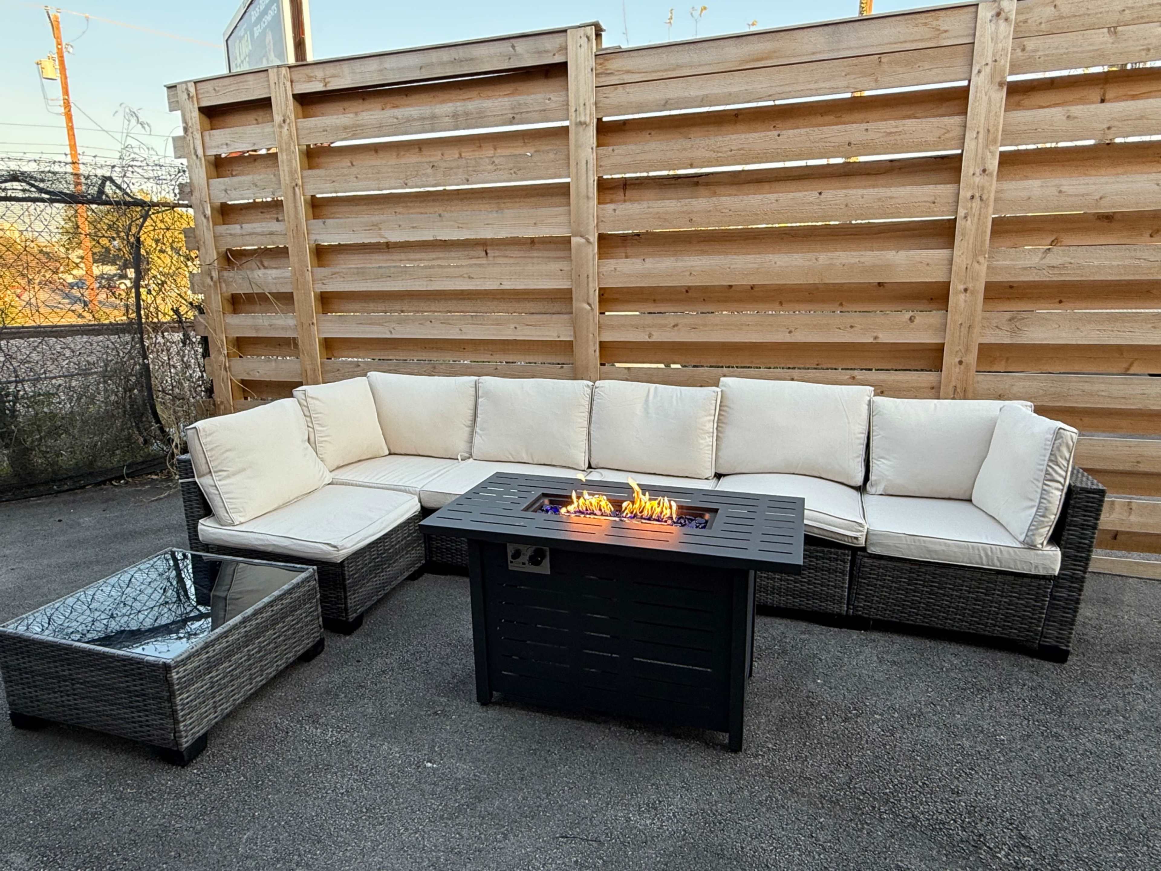 The image shows a cushioned sectional sofa and a rectangular fire pit with flames, set on a concrete surface enclosed by a wooden slatted fence.