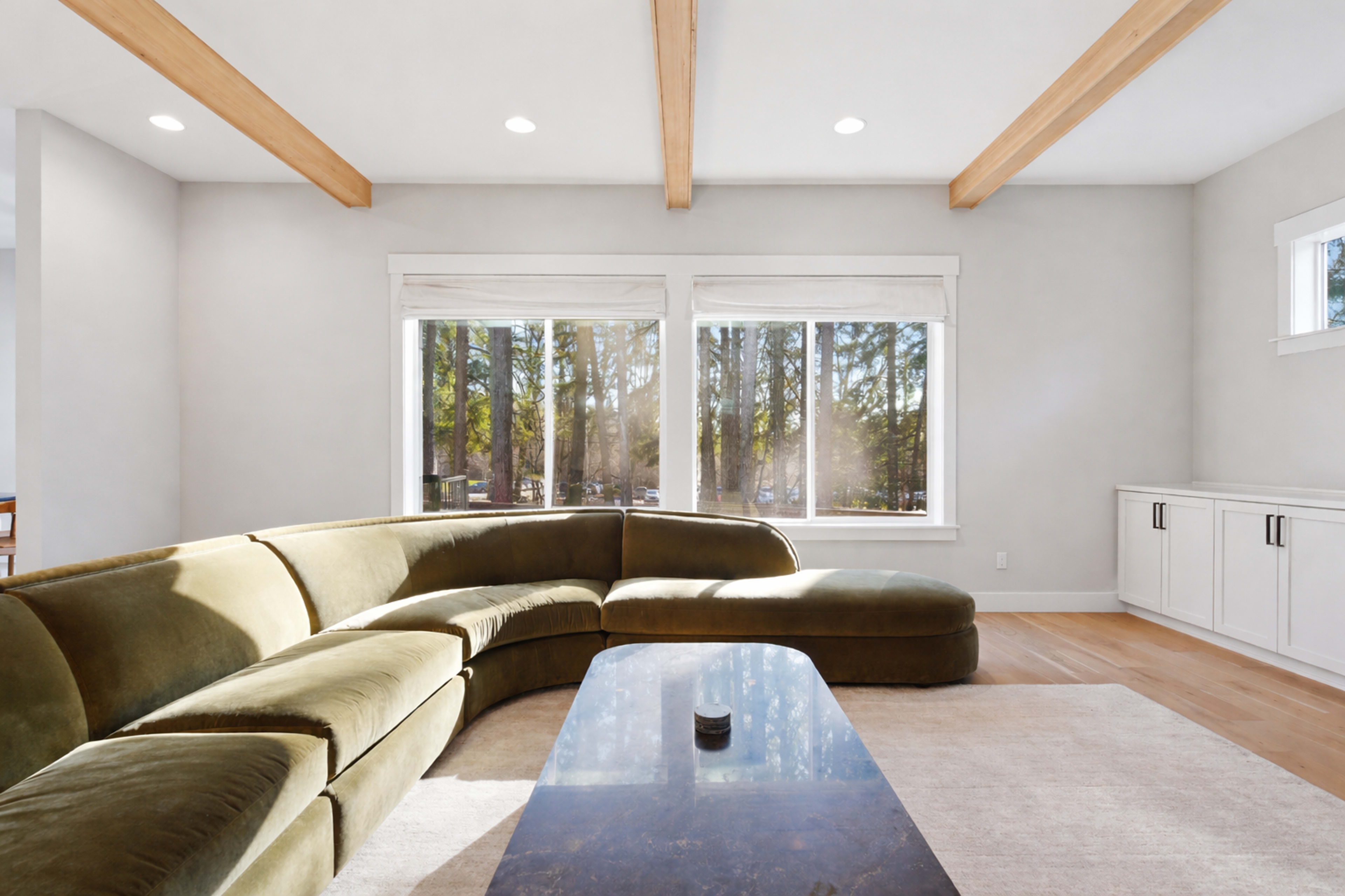The image shows a modern living room with a curved green sofa, a dark coffee table, and large windows revealing a view of trees outside.