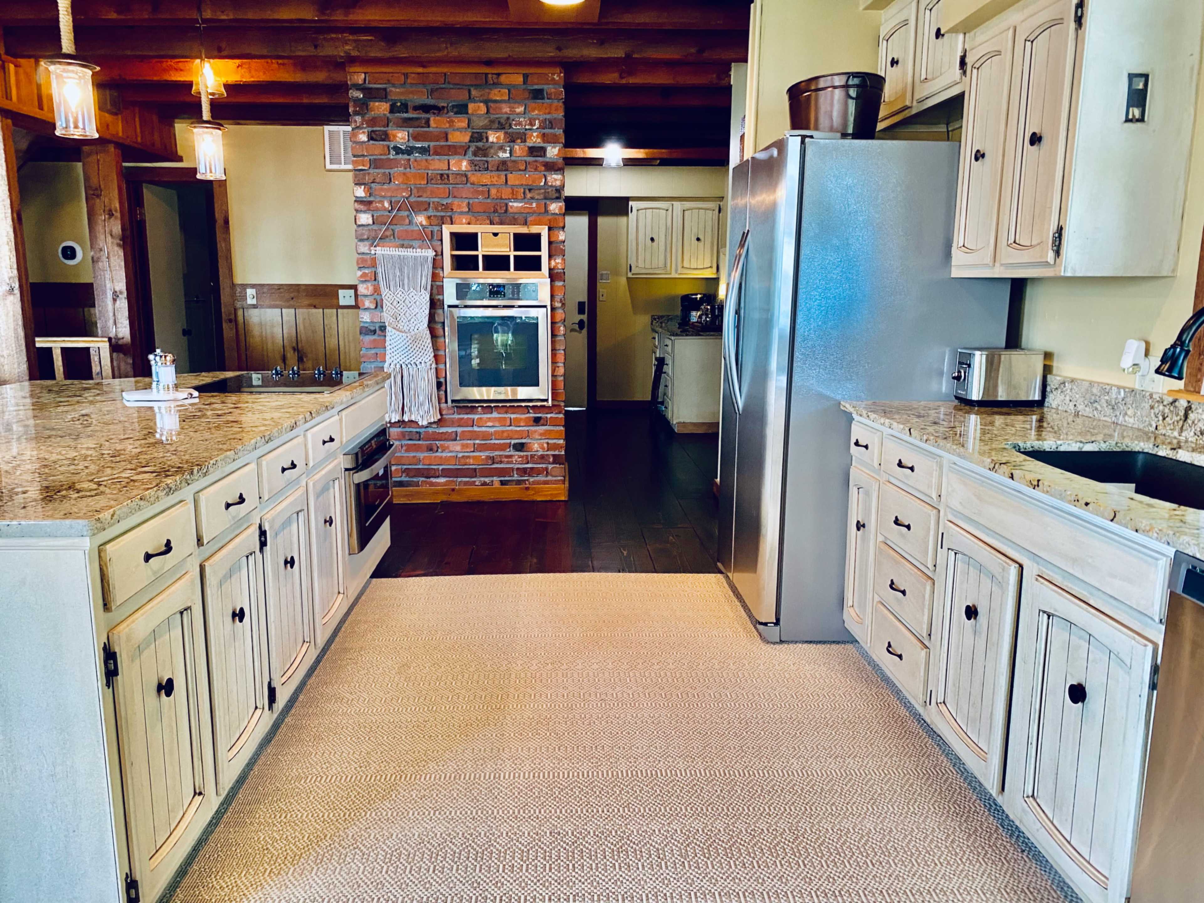 The image shows a kitchen with a central island, granite countertops, wooden beams on the ceiling, and a brick accent wall featuring an oven and a stainless-steel refrigerator.