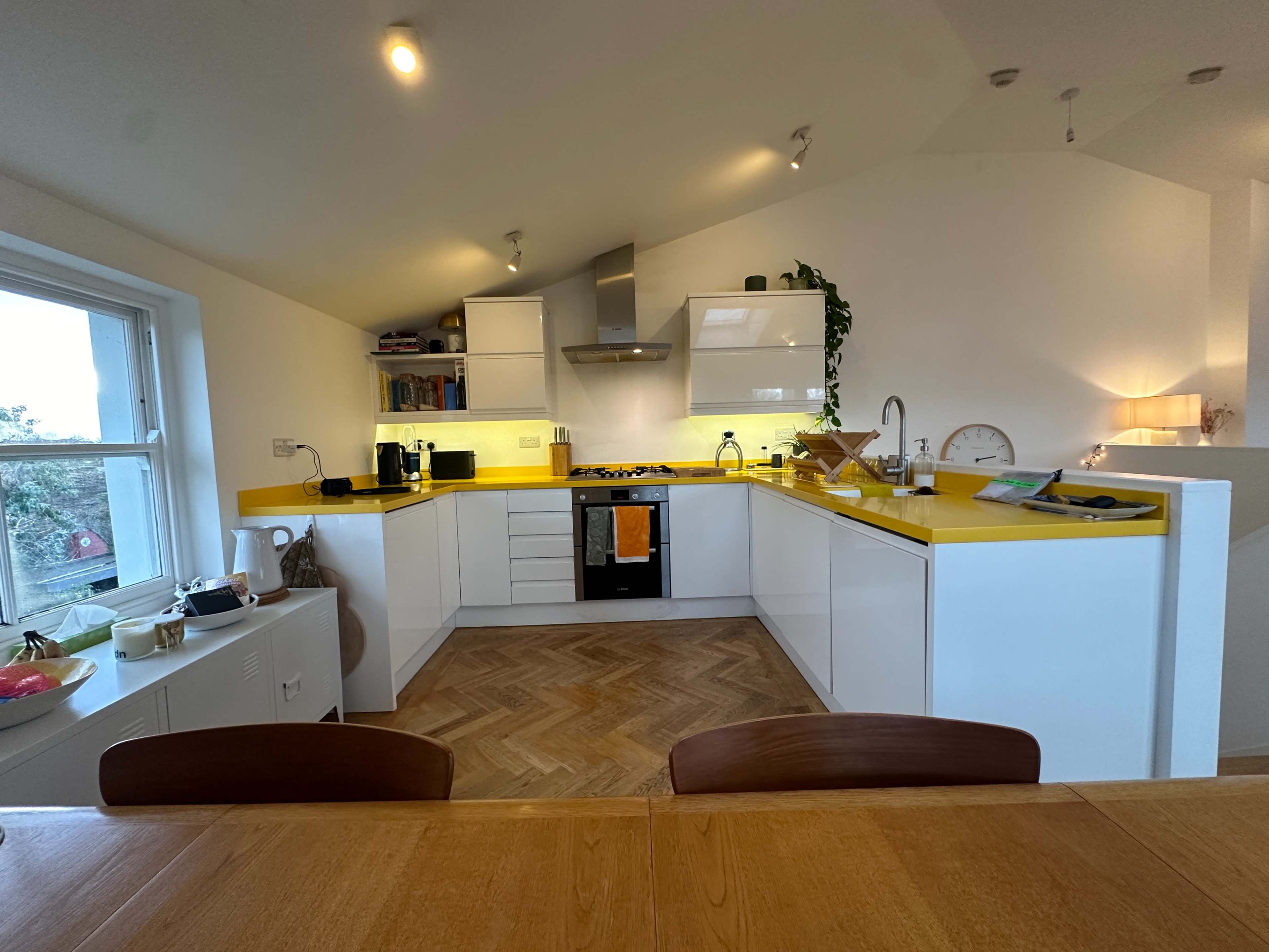The image shows a modern kitchen with yellow countertops, white cabinets, and a herringbone pattern wooden floor.