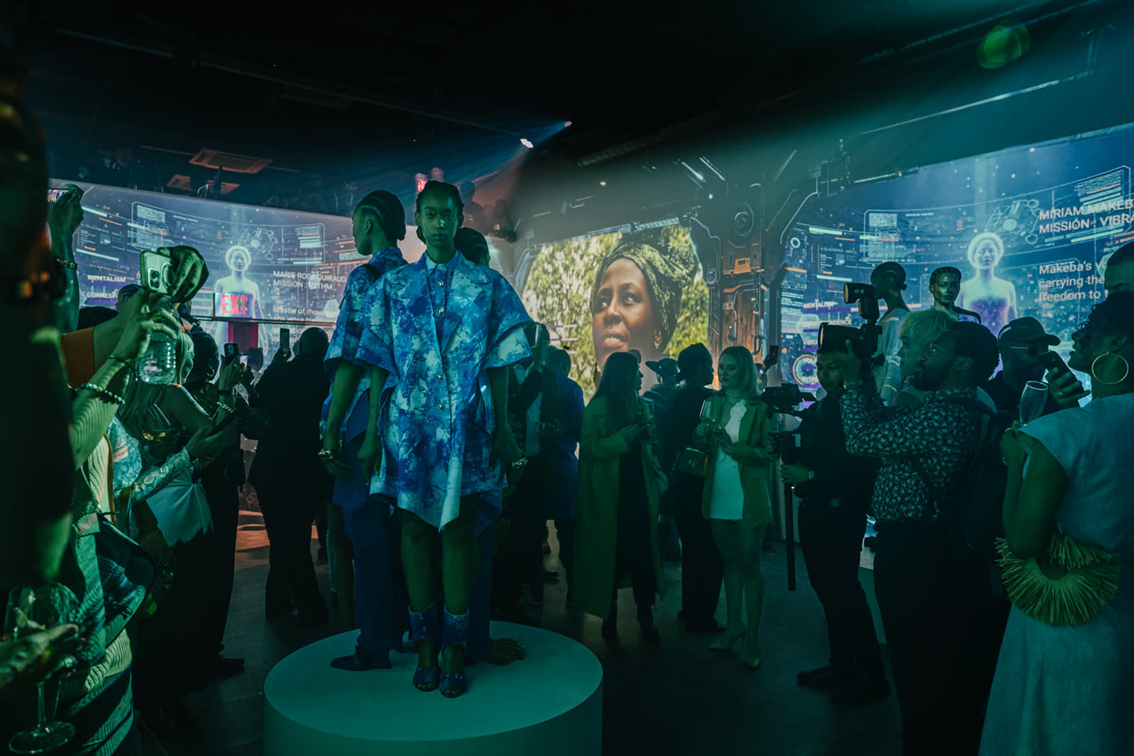 A crowd of people mingles in a vibrant event space, featuring a central model on a podium and large screens displaying digital visuals.