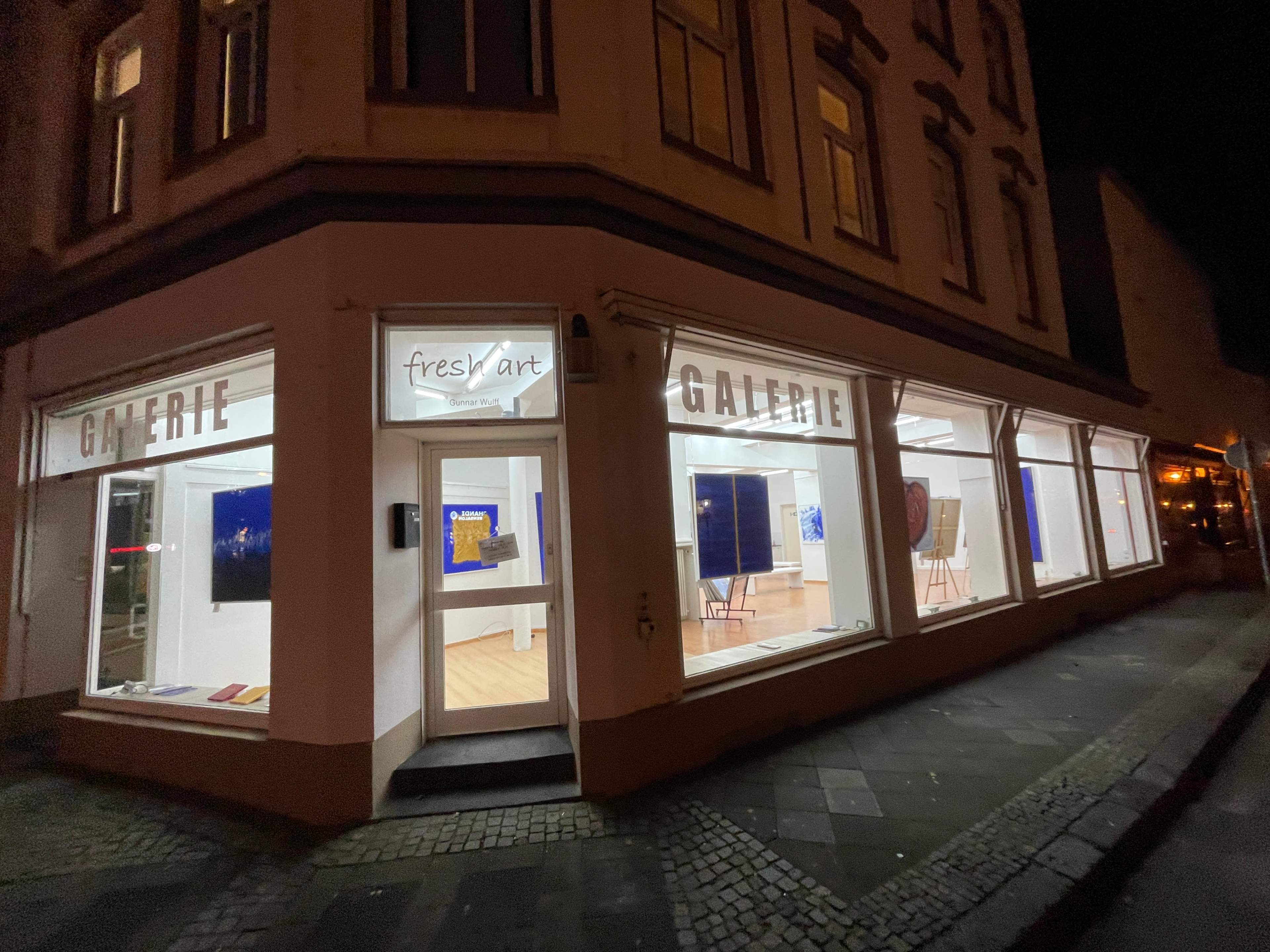 A lit art gallery at night, featuring large windows displaying various artworks.