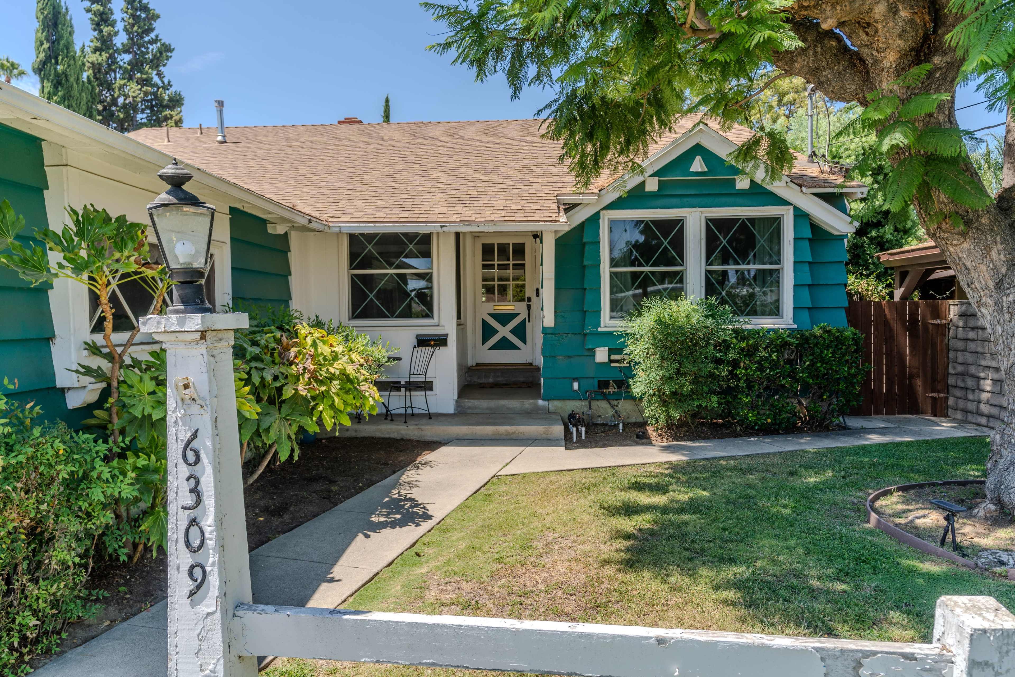 The image shows a blue and green house with a front porch, surrounded by a lawn and shrubs.